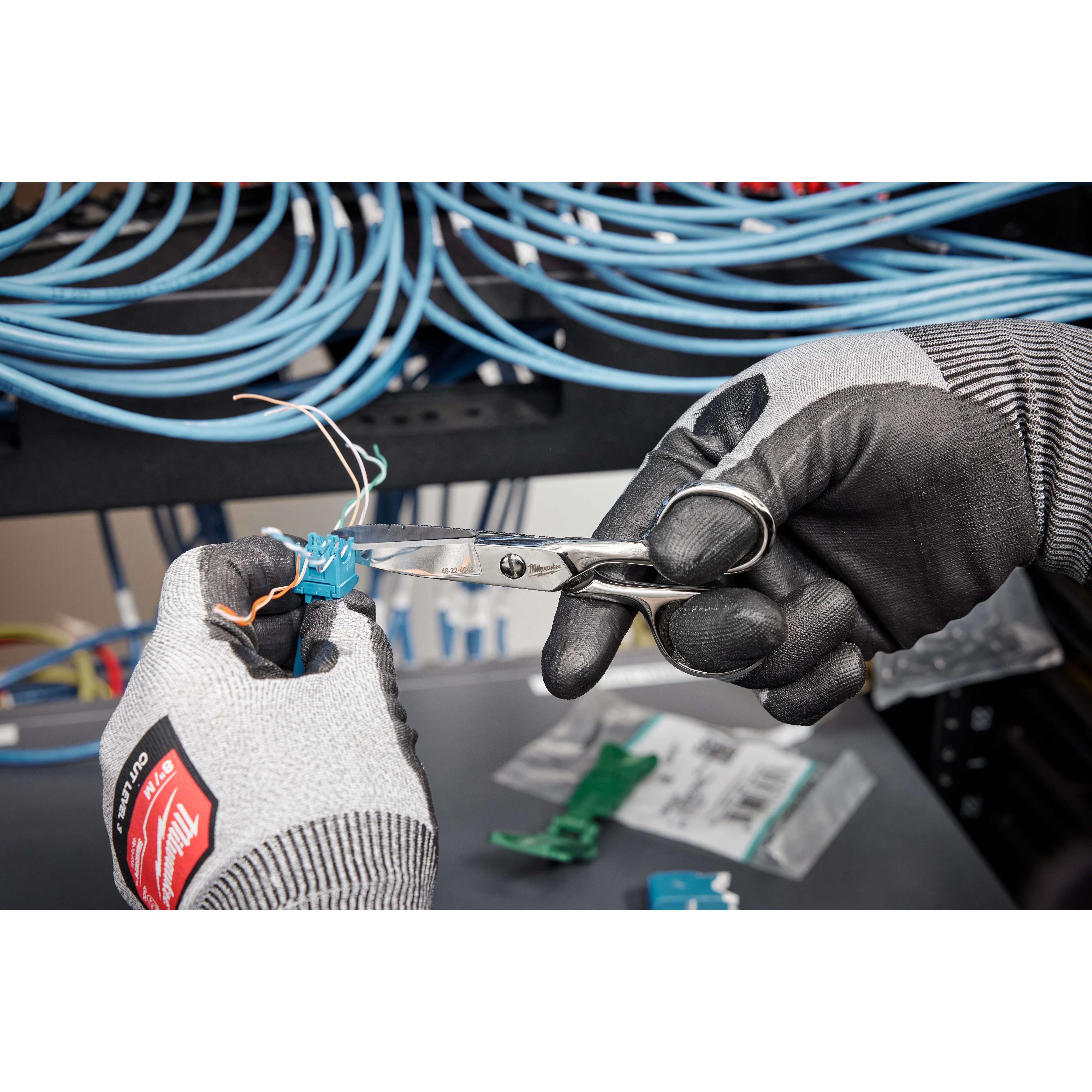 Hands wearing black gloves use Electrician Scissors to cut wires from a blue cable. Coiled blue cables are visible in the background.
