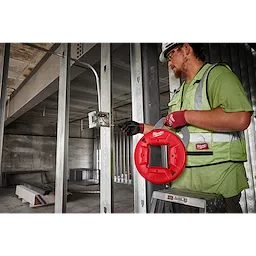 A person using a 120' 1/8" Stainless Steel Fish Tape at a construction site. The person is wearing gloves and a green safety vest.