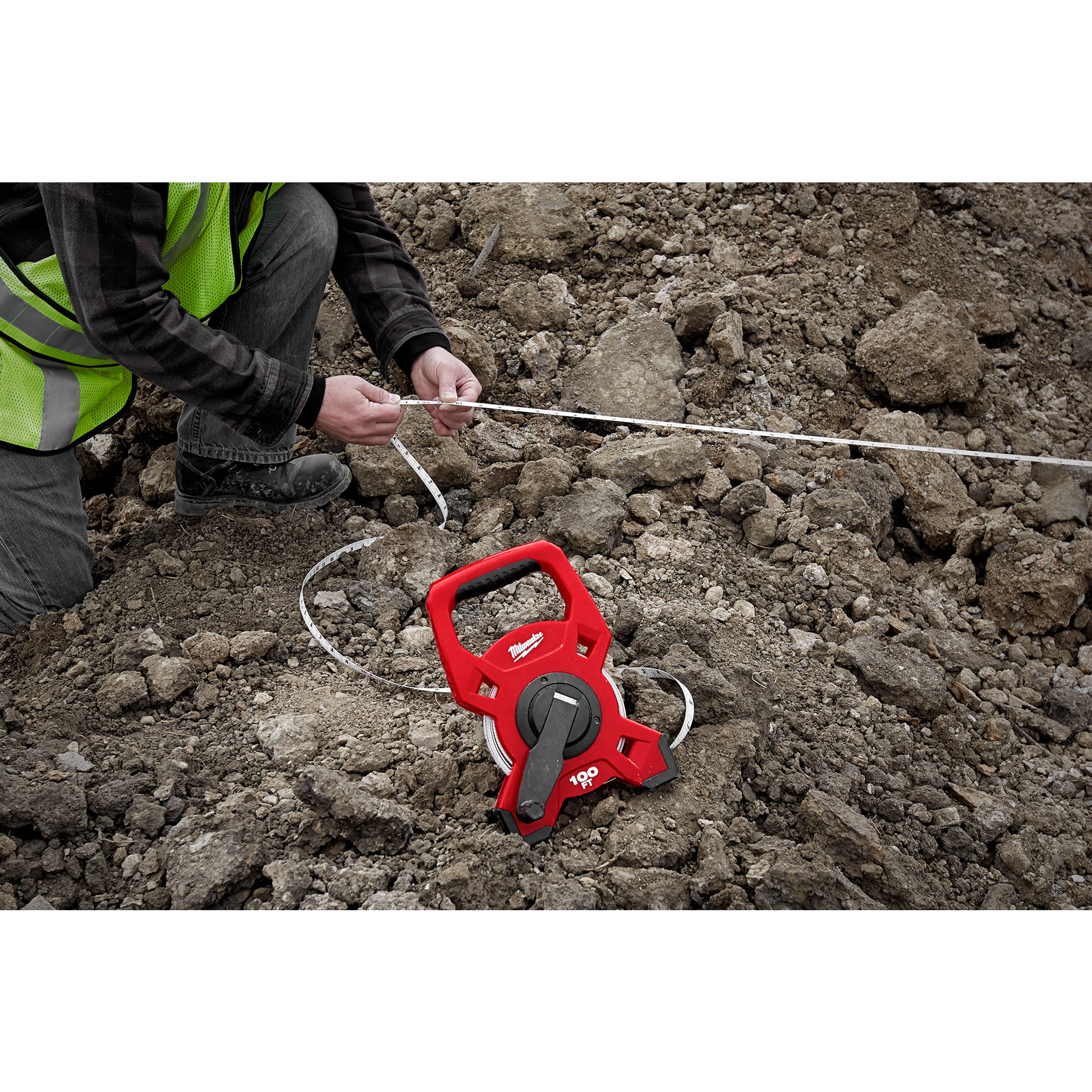 Person using a Milwaukee 100 Foot Fiberglass Long Tape on rocky terrain, with the red tape measure tool spread out.