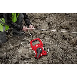 Person using a Milwaukee 100 Foot Fiberglass Long Tape on rocky terrain, with the red tape measure tool spread out.