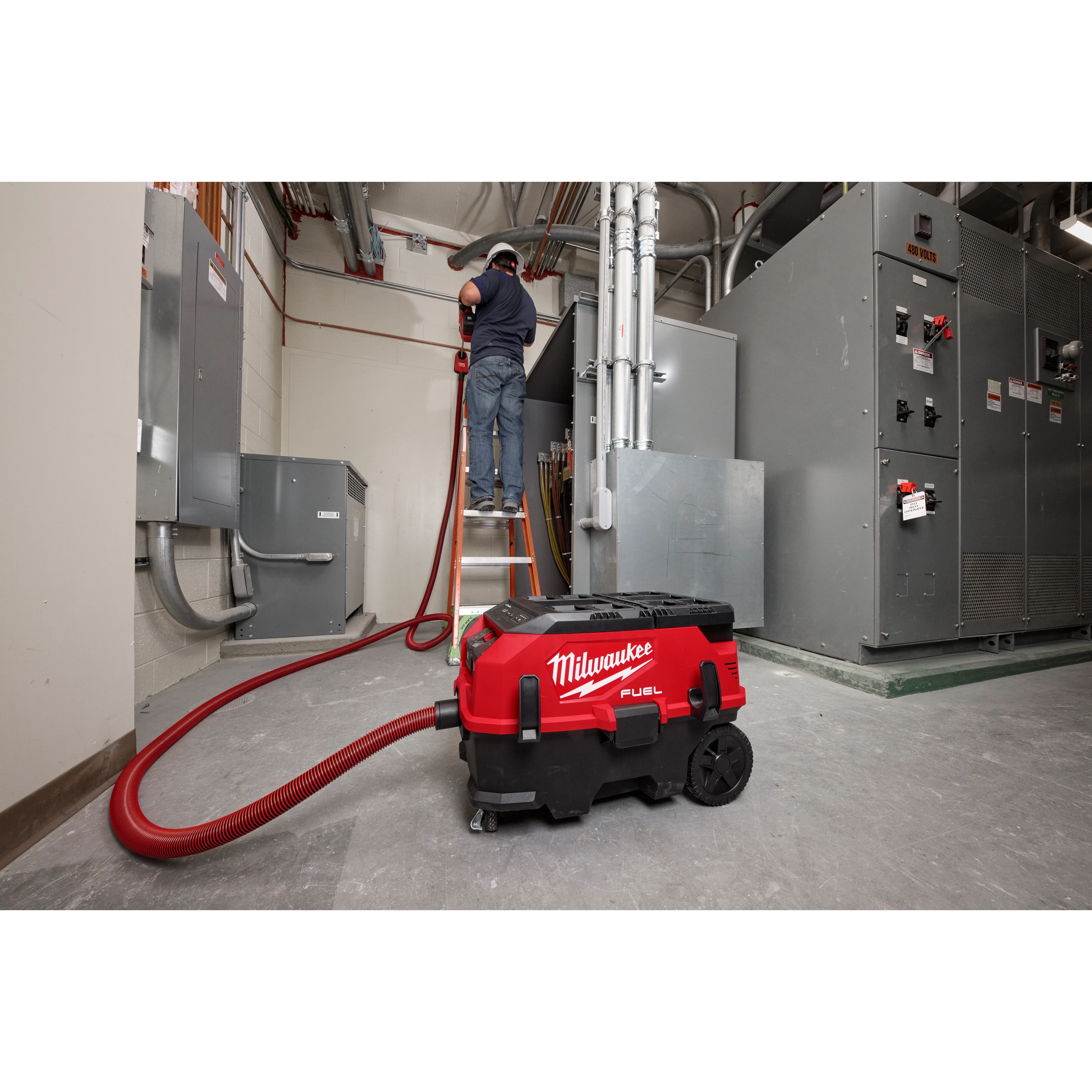 A worker is seen using a red and black Milwaukee M18 FUEL™ PACKOUT™ 9 Gallon Dual-Battery Dust Extractor with VACLINK™ in an industrial setting. The dust extractor is on the floor with its hose extended and attached, while the worker is elevated on a ladder.
