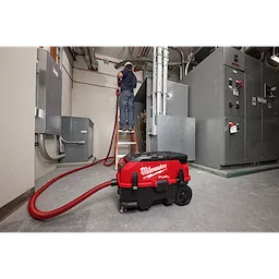 A worker is seen using a red and black Milwaukee M18 FUEL™ PACKOUT™ 9 Gallon Dual-Battery Dust Extractor with VACLINK™ in an industrial setting. The dust extractor is on the floor with its hose extended and attached, while the worker is elevated on a ladder.