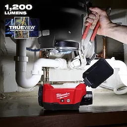 Milwaukee M12™ ROVER™ Multi-Directional Flood Light w/ PACKOUT™ Compatibility is shown illuminating plumbing work under a sink. The red light features TrueView high definition output and delivers 1,200 lumens. A hand is using pliers to work on pipes, benefiting from the versatile light direction.