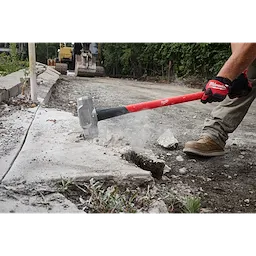 A construction worker uses a 10lb Sledge Hammer (36" Handle) to break apart concrete on a worksite. The sledgehammer has a red handle and its metal head impacts the pavement, causing debris and dust to scatter. The worker wears gloves and sturdy boots, standing on gravel next to heavy machinery.