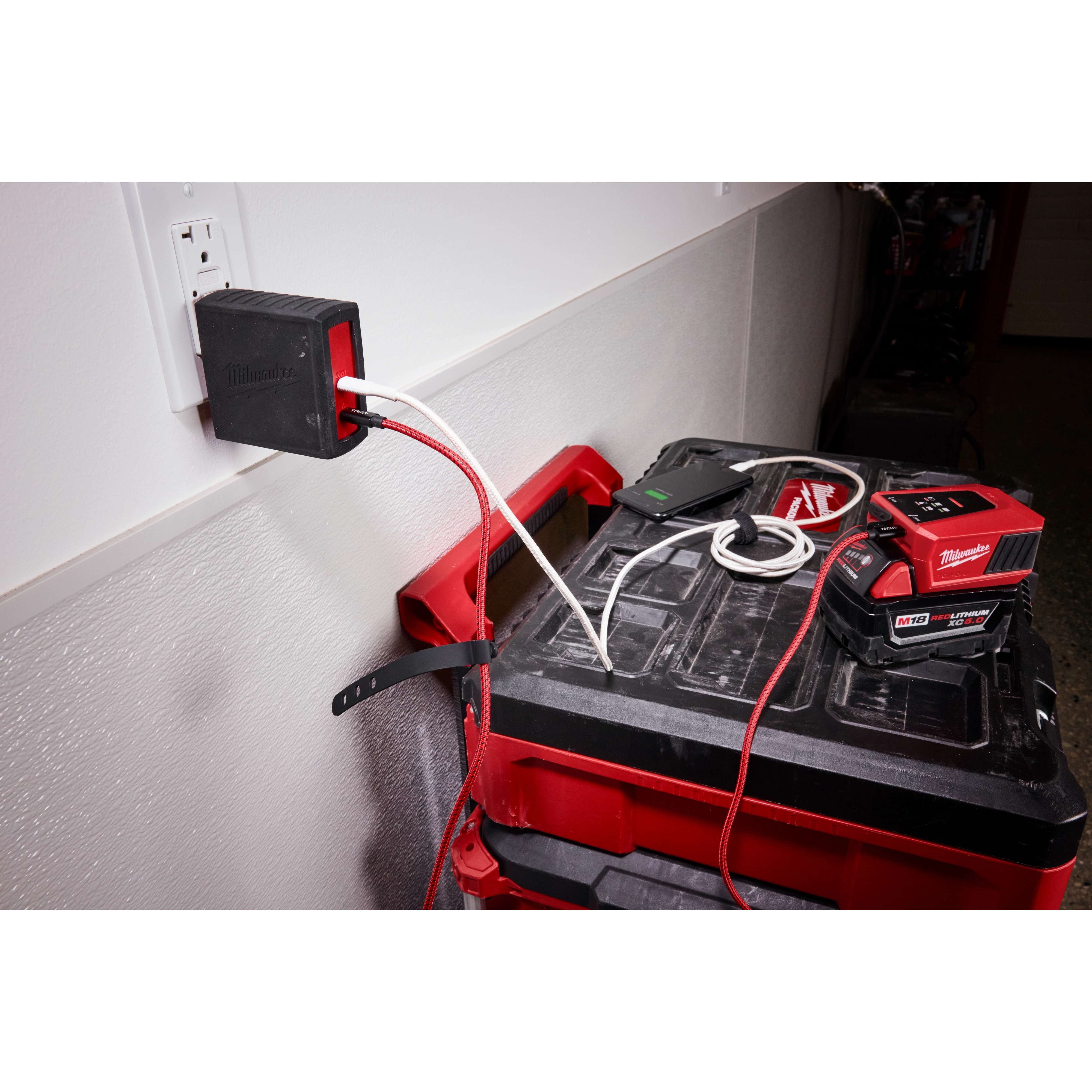 The image displays the Milwaukee 120W USB-C 2-Port Wall Adapter plugged into a wall socket, charging multiple devices.