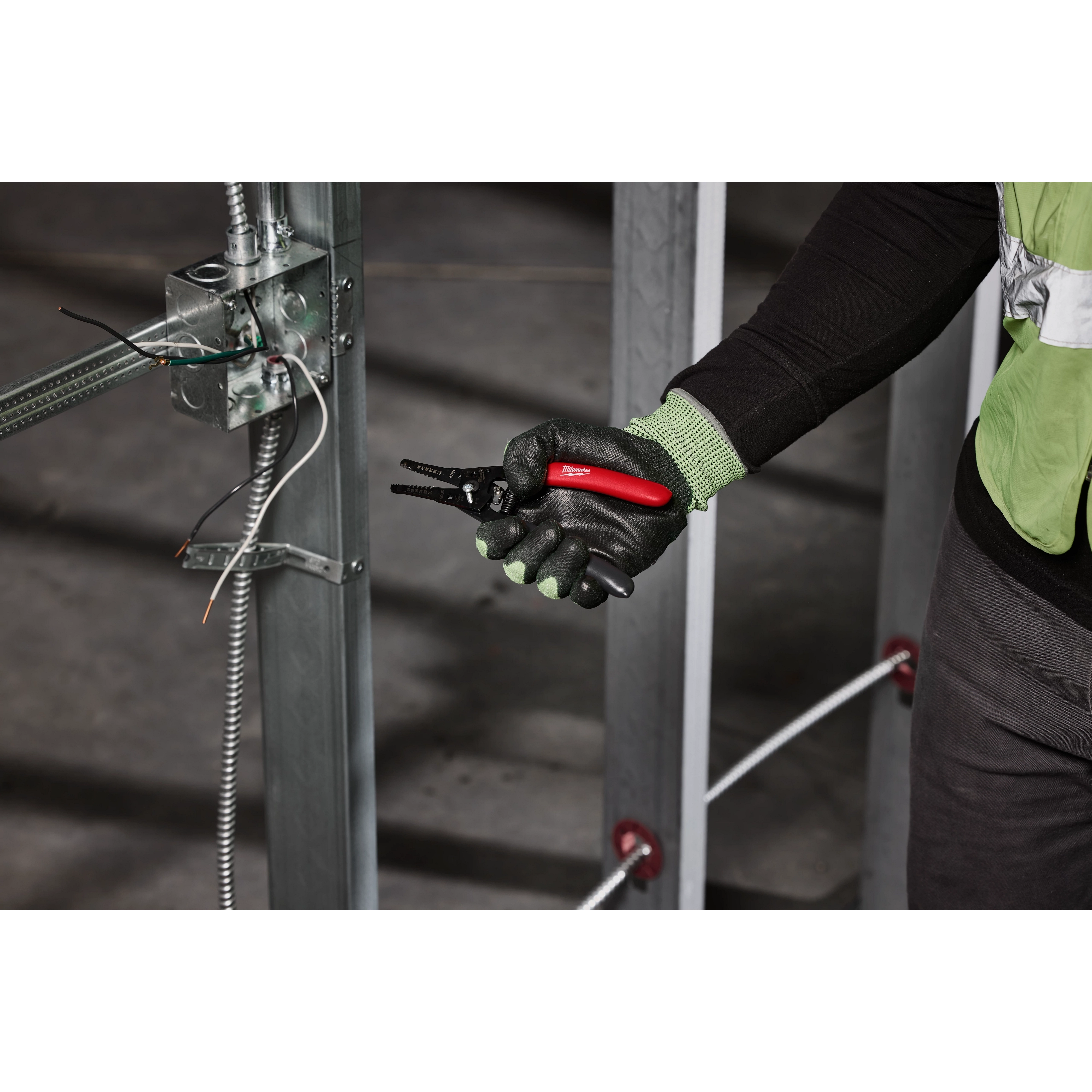 A person wearing gloves holds a 20-32 AWG Low Voltage Dipped Grip Wire Stripper & Cutter. The person is working on an electrical panel with wires.
