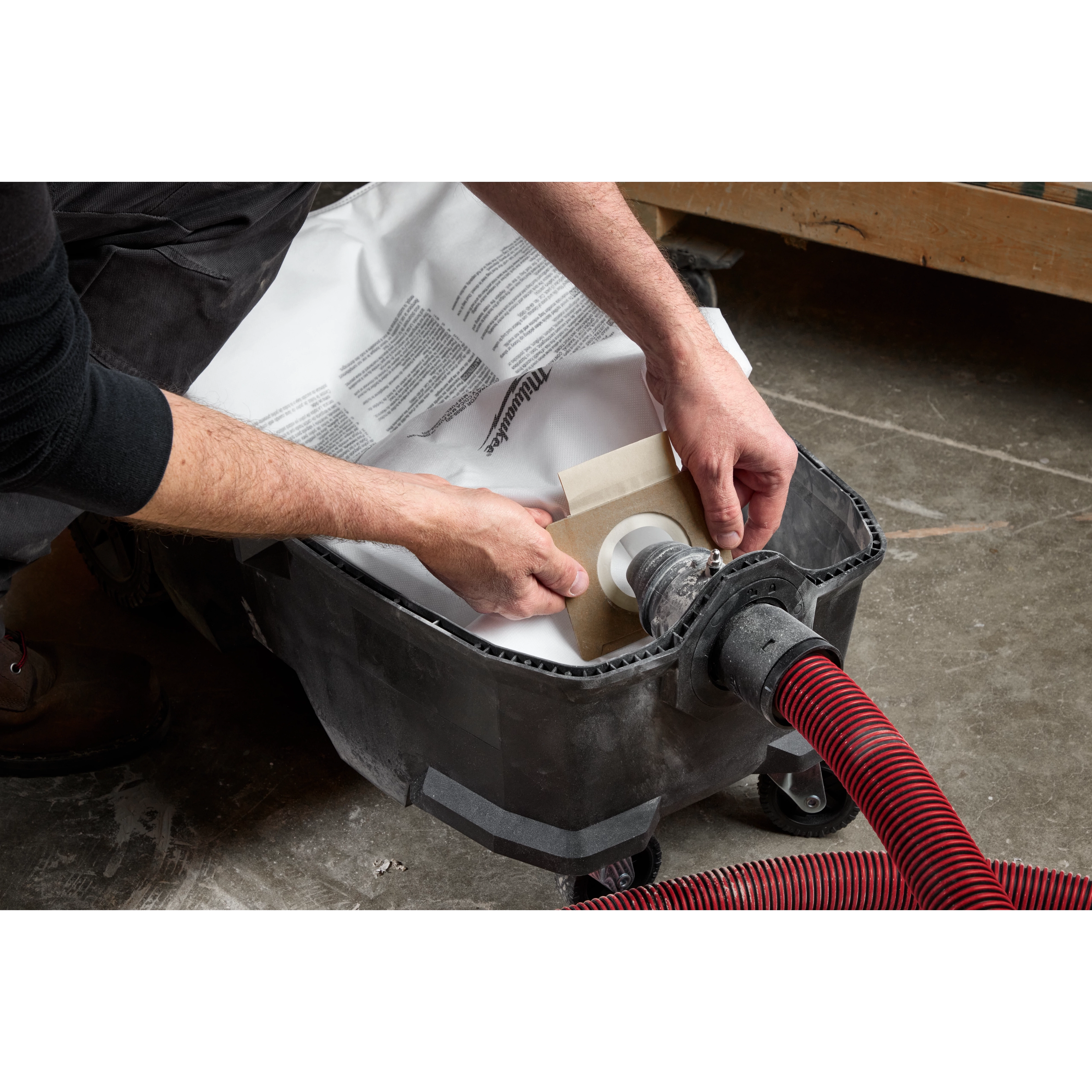 A person is installing one of the Fleece Dust Bags (5PK) into a vacuum cleaner. The vacuum has a red and black hose attached, and the person's hands are placing the bag into the compartment.