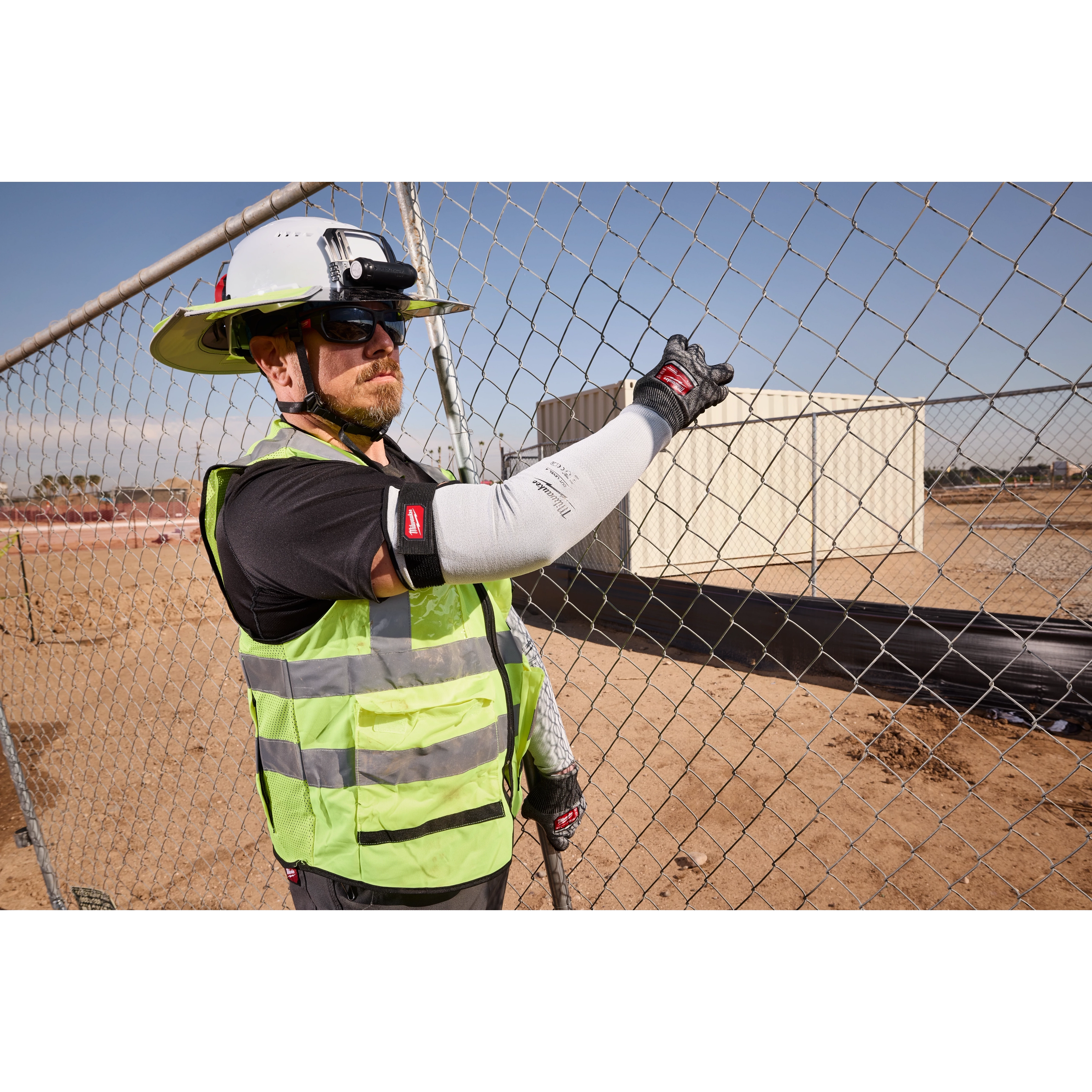 Worker wearing Cut Level 4 Protective Sleeves and high-visibility vest, handling a chain-link fence at a construction site.