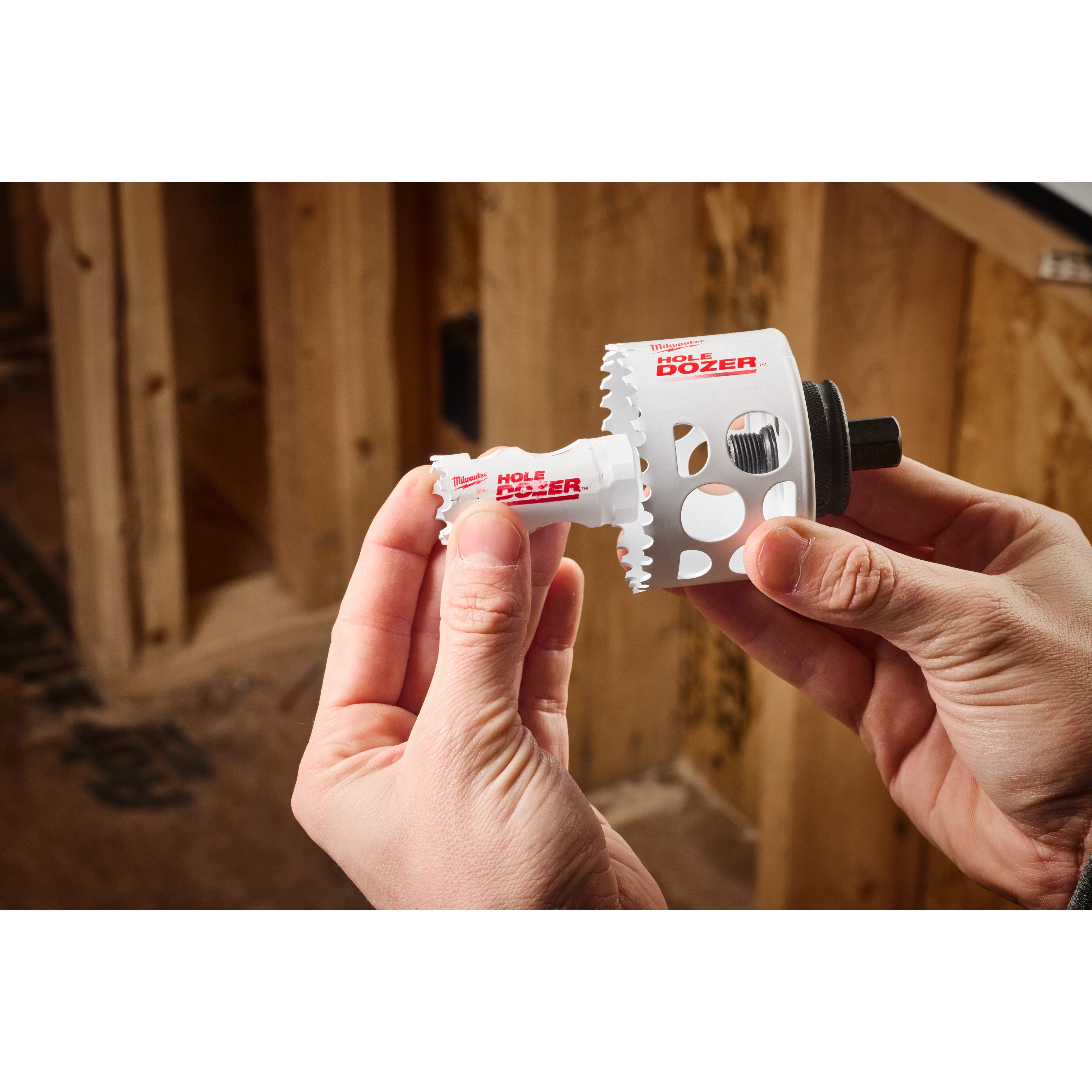 A person holds a Hole Enlargement Adapter for QUIK-LOK™ Arbor tool by Milwaukee in a woodworking shop. The adapter is white with serrated edges and labeled "Hole Dozer."