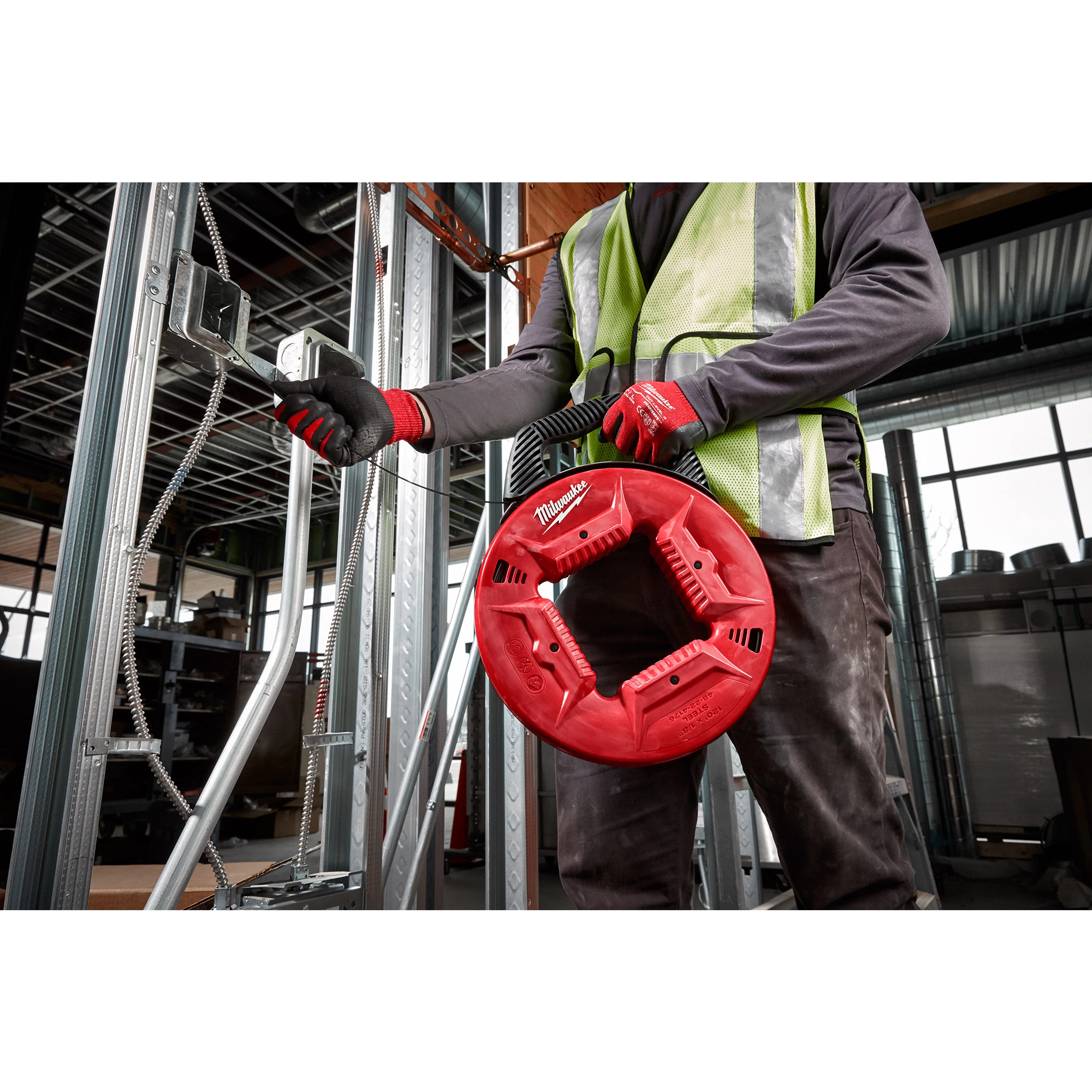 Worker holding a red 1/8" 120' Steel Fish Tape, threading it through metal electrical conduit in a construction setting.