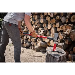A person uses an 8lb Splitting Maul (36" Handle) to split a large piece of wood. The maul features a grey and red handle and is shown in interaction with a log. There are stacks of cut logs in the background. The person is wearing grey pants and a light shirt.
