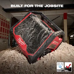 The M18 BLUETOOTH JOBSITE RADIO + CHARGER is shown on a dusty construction site. The rugged, red and black device features dust and water resistance (IP65) and impact resistance. The product is designed for durability and tough work environments, enhancing the job site audio experience while charging tools.