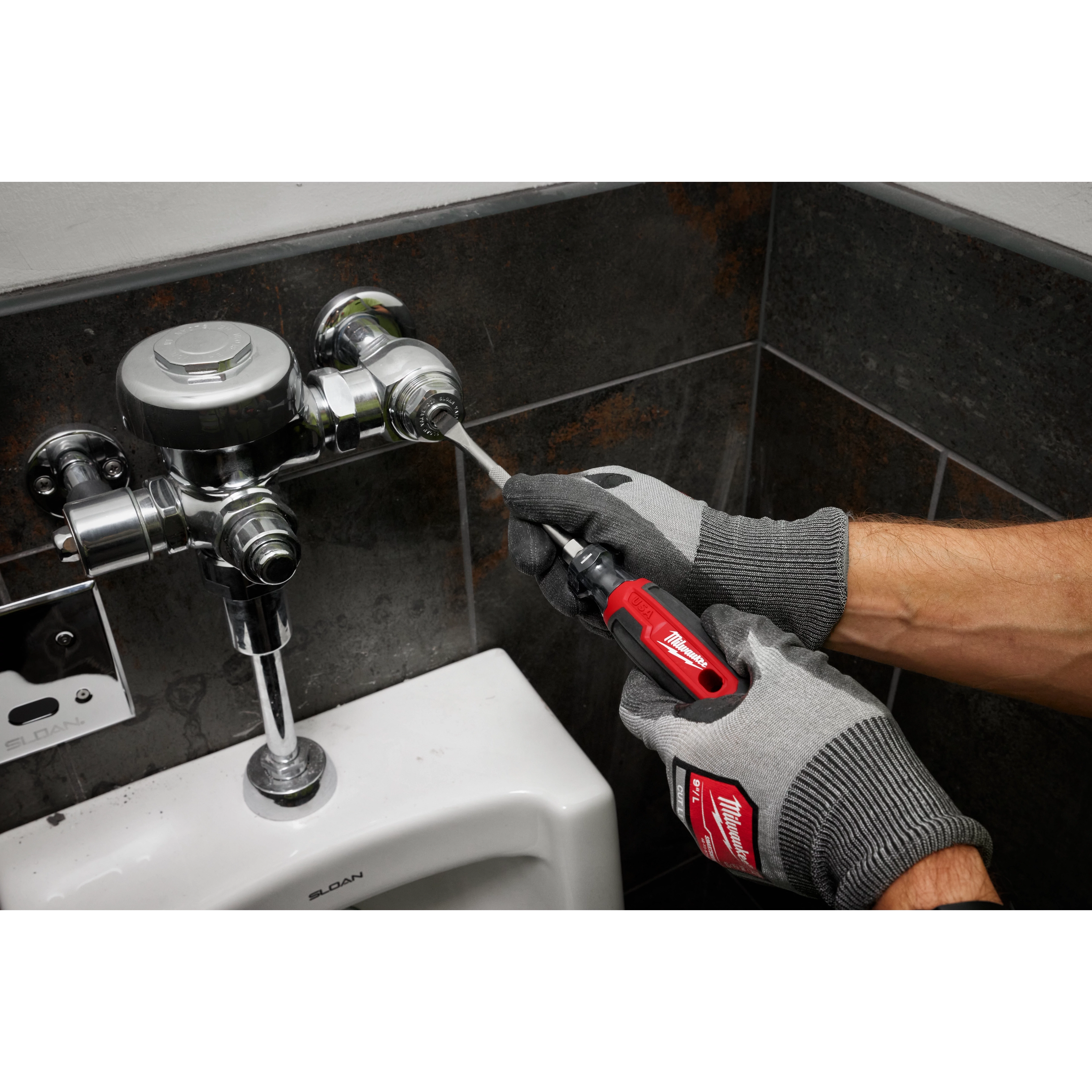 A person wearing gloves uses a 5/16" Slotted 6" Cushion Grip Screwdriver (USA) to adjust a plumbing fixture above a urinal.