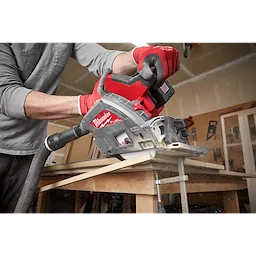 A person is using a Milwaukee M18 FUEL 6 1/2" Plunge Track Saw on a piece of wood in a workshop, creating sawdust.