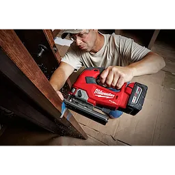 A person is using an M18 FUEL Jigsaw, D-Handle to cut through a wooden surface. The tool is red with a black base.