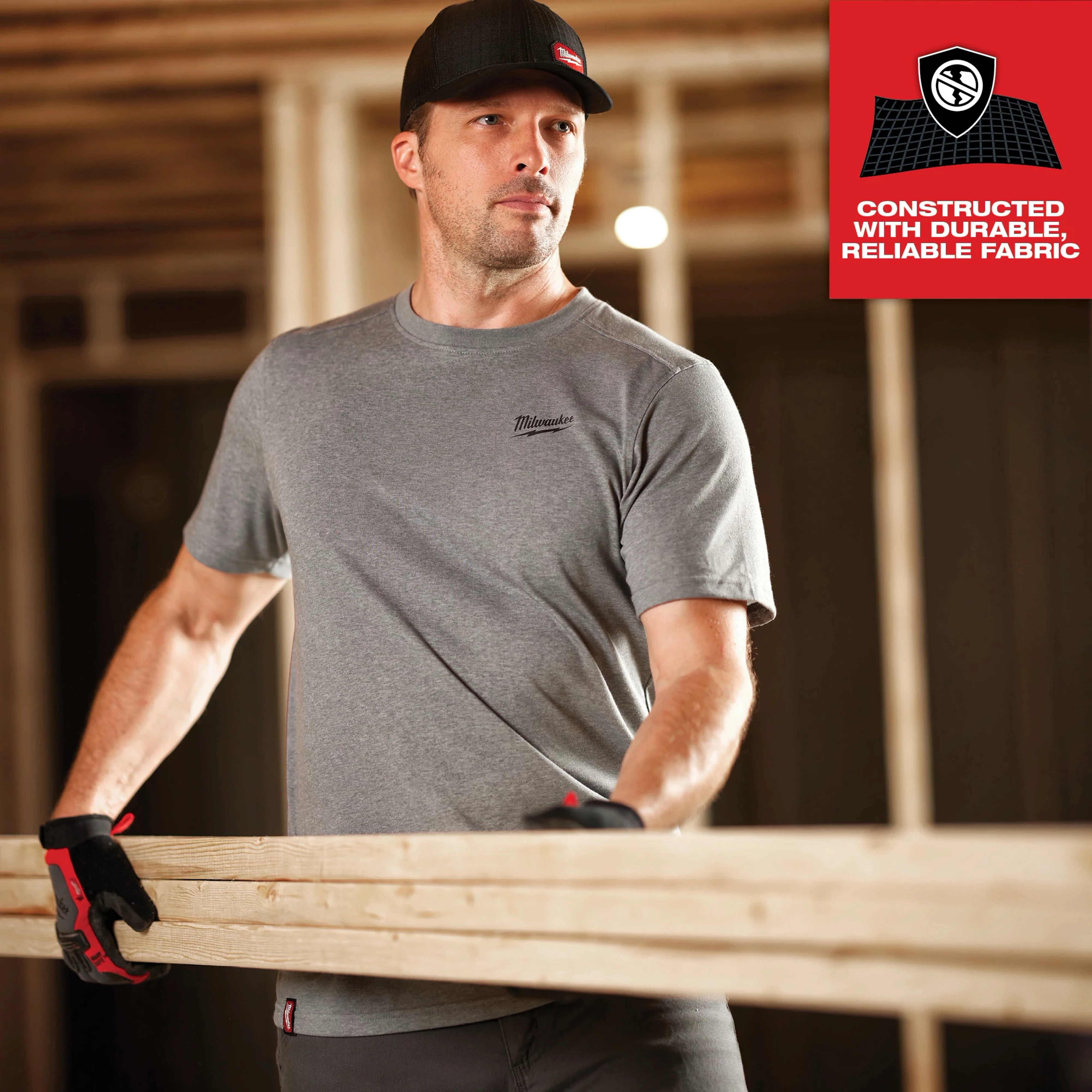 Man wearing GRIDIRON Snapback Trucker Hat - Black, gray t-shirt, and work gloves, holding a wooden plank at a construction site.