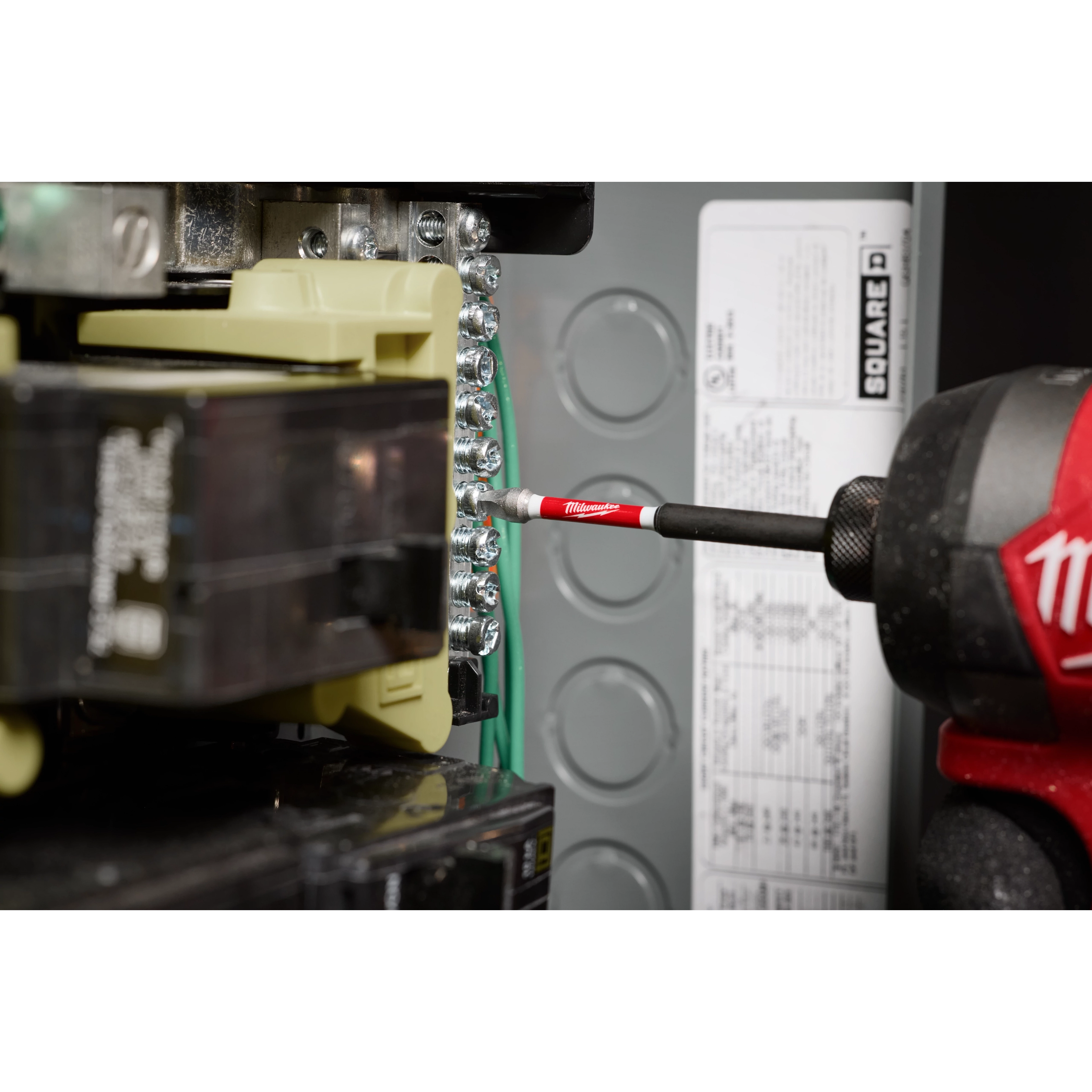 A SHOCKWAVE™ 3-1/2” Impact Square Recess #2 Power Bit is shown in use, attached to a power drill, working on an electrical panel. The bit is red and black with the brand name visible, and it is being used to tighten screws inside the panel.