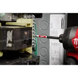 A SHOCKWAVE™ 3-1/2” Impact Square Recess #2 Power Bit is shown in use, attached to a power drill, working on an electrical panel. The bit is red and black with the brand name visible, and it is being used to tighten screws inside the panel.