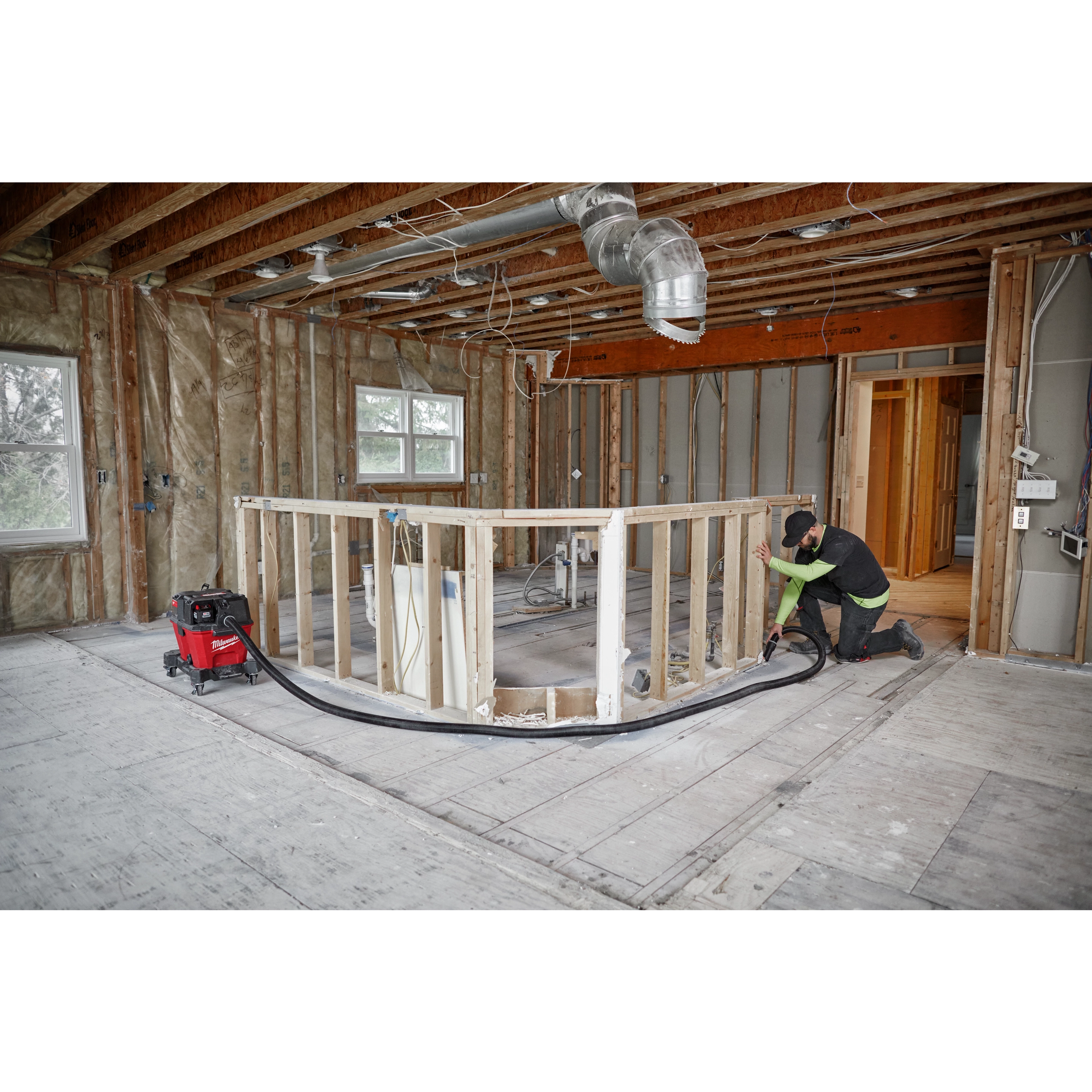 A worker using a 1-7/8” x 16' flexible hose connected to a red vacuum in a partially constructed home interior.