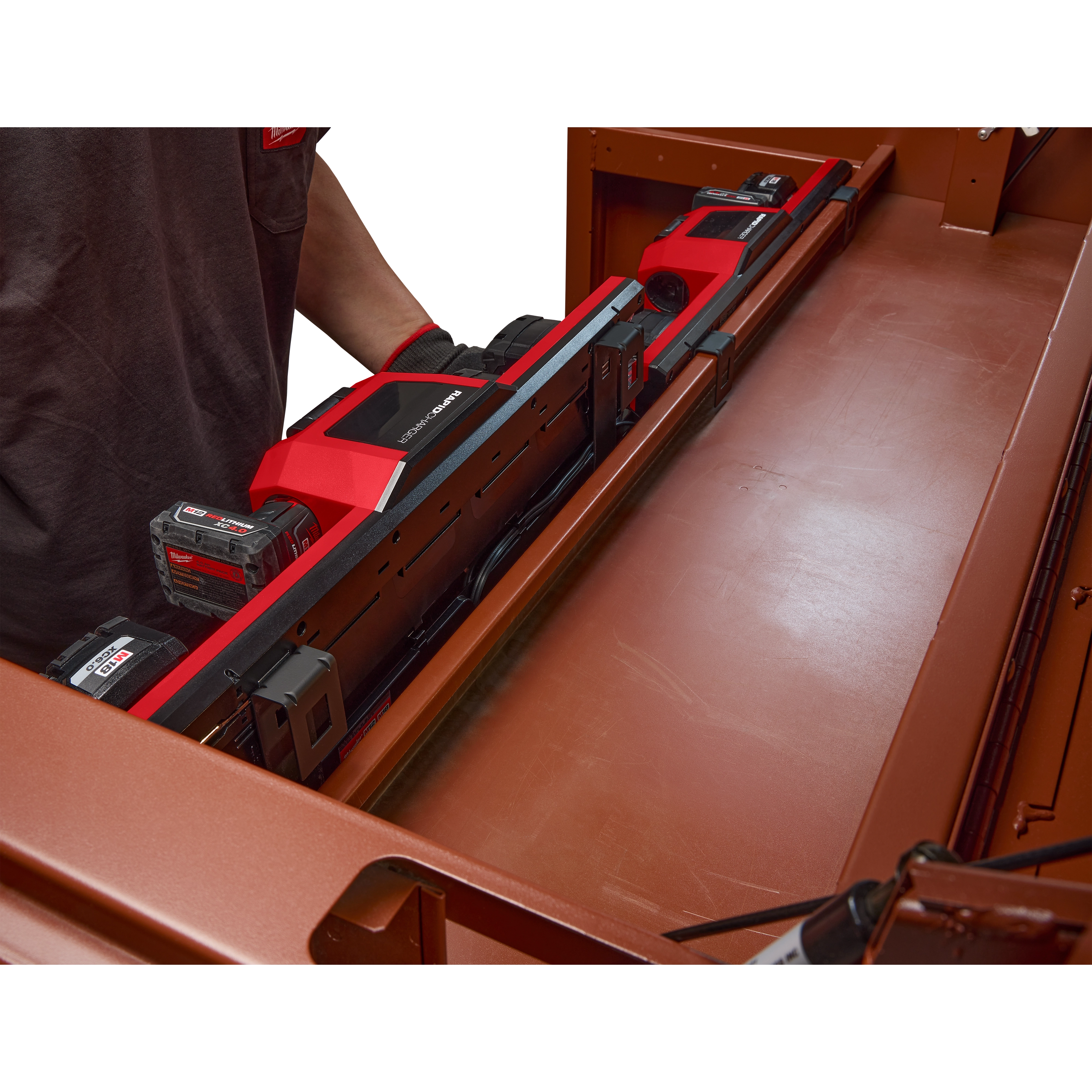 The image shows an M18™ and M12™ Gangbox Rapid Charger inside a metal toolbox. The charger is designed to charge multiple batteries simultaneously and is seen holding seven battery packs. A person wearing a gray shirt and gloves is placing or removing the charger from the box.