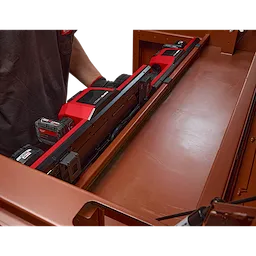 The image shows an M18™ and M12™ Gangbox Rapid Charger inside a metal toolbox. The charger is designed to charge multiple batteries simultaneously and is seen holding seven battery packs. A person wearing a gray shirt and gloves is placing or removing the charger from the box.