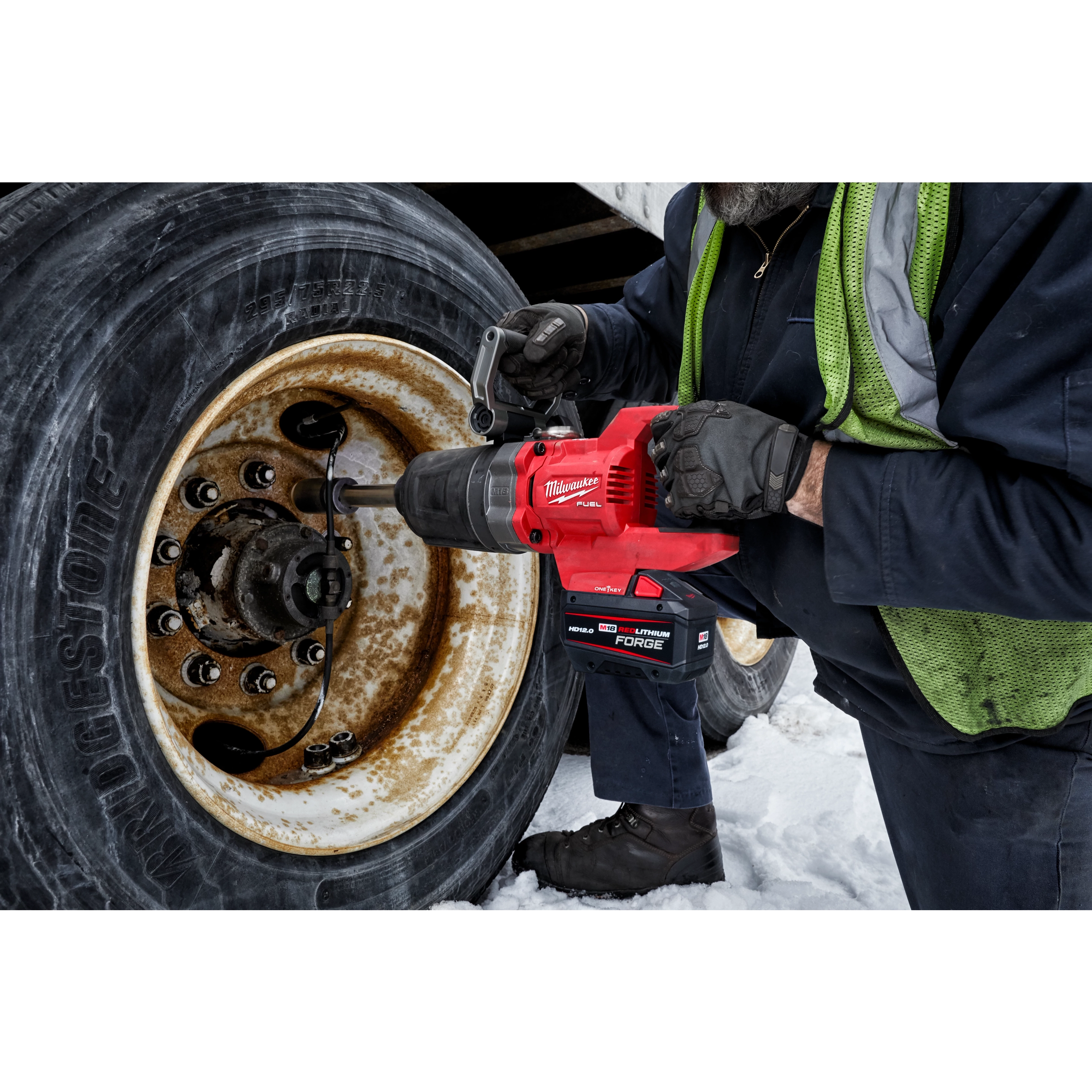 A worker operates an M18 FUEL™ 1" D-Handle Extended Anvil High Torque Impact Wrench w/ONE-KEY™ App Shot to loosen the bolts on a large truck tire. Snow is visible on the ground beneath the truck.