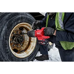 A worker operates an M18 FUEL™ 1" D-Handle Extended Anvil High Torque Impact Wrench w/ONE-KEY™ App Shot to loosen the bolts on a large truck tire. Snow is visible on the ground beneath the truck.