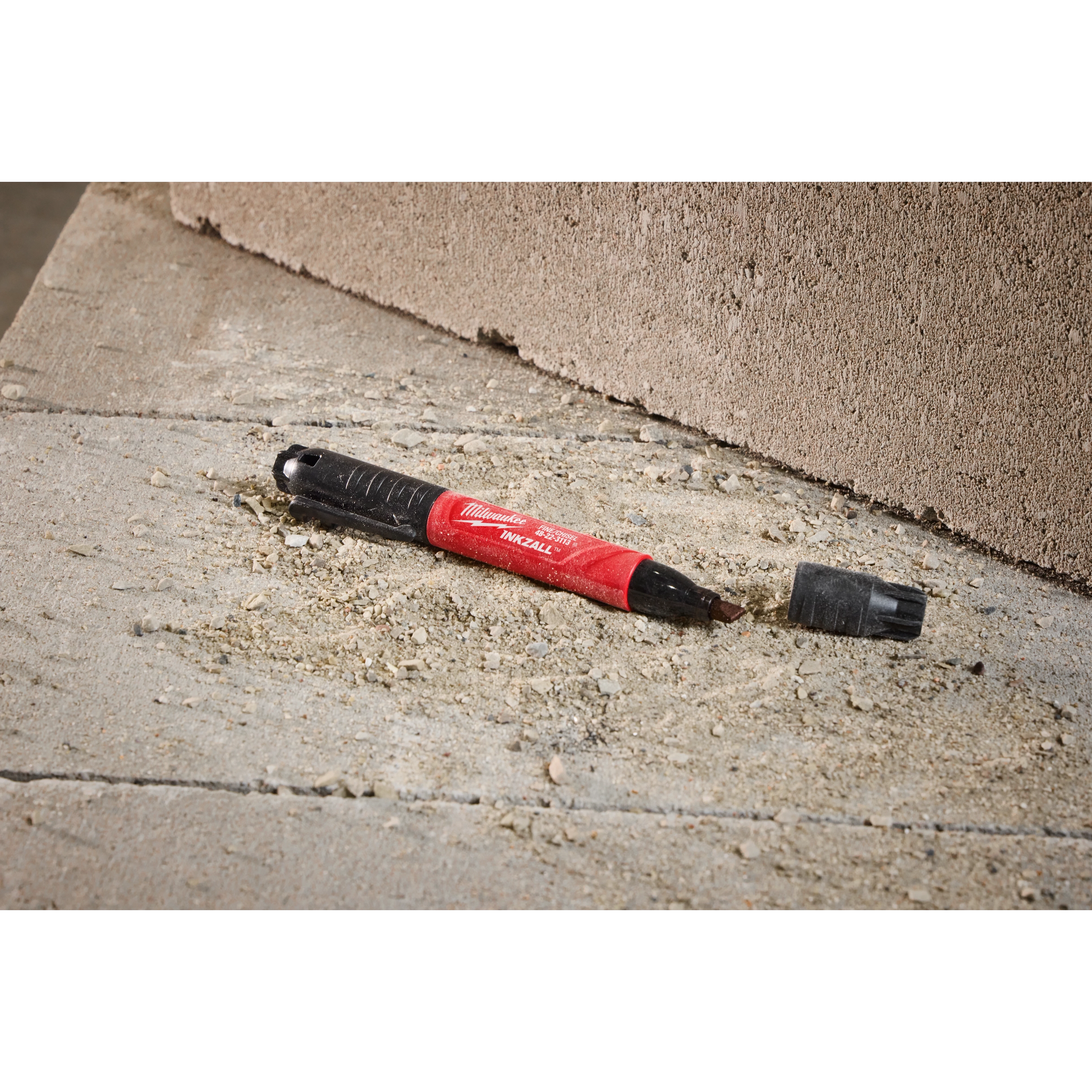 The INKZALL Double-Sided Black Jobsite Marker has a Fine Point & Chisel Tip. It rests on a rough, sandy concrete surface. The red and black marker has its cap off and lies next to an unfinished block wall.
