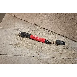 The INKZALL Double-Sided Black Jobsite Marker has a Fine Point & Chisel Tip. It rests on a rough, sandy concrete surface. The red and black marker has its cap off and lies next to an unfinished block wall.