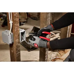 A person wearing gloves uses a 10-28 AWG Multi-Purpose Dipped Grip Wire Stripper & Cutter with Reinforced Head to strip wires in an electrical outlet box on a wooden stud.