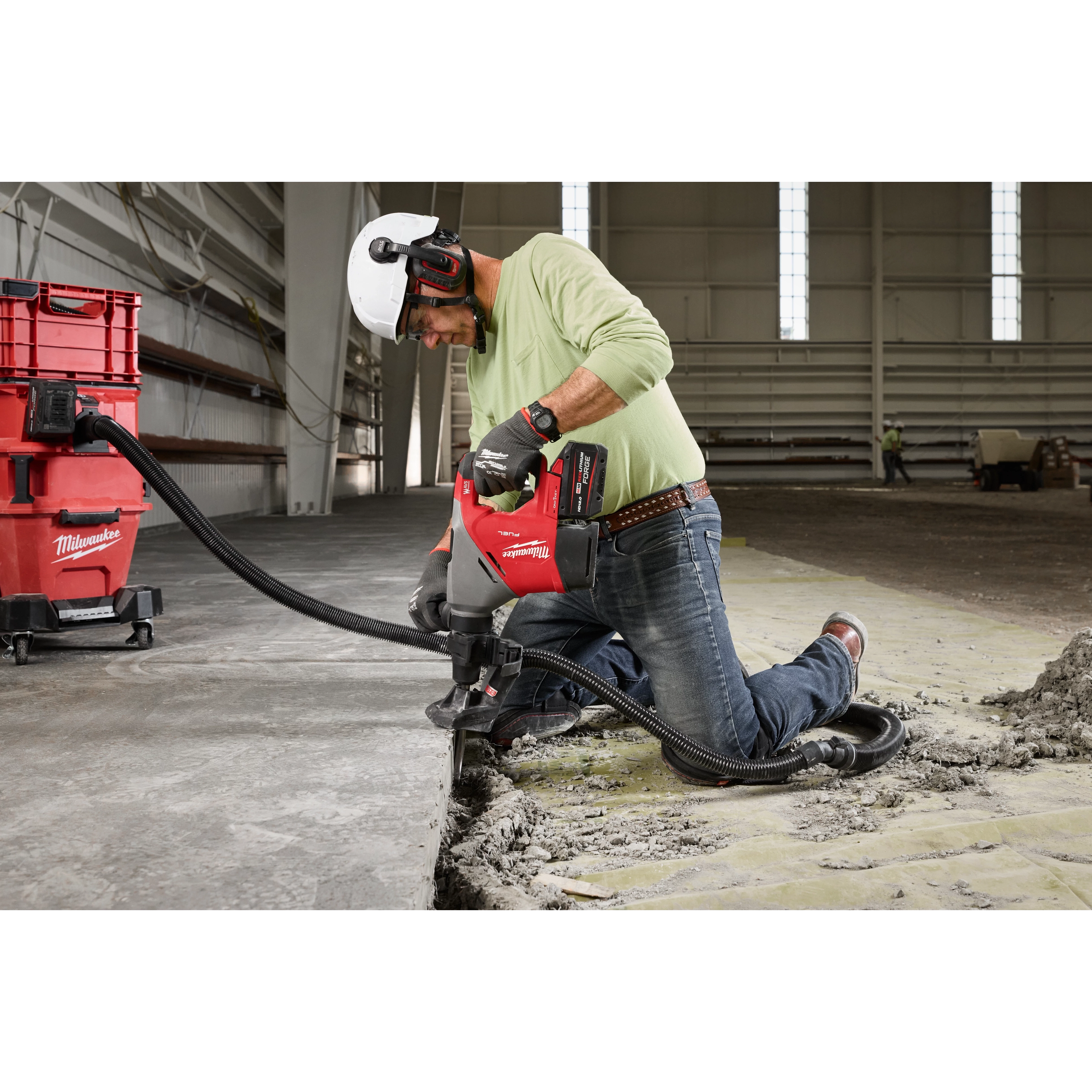 A worker in a construction site uses the M18™ FUEL™ 1-9/16" SDS Max Rotary Hammer w/ ONE-KEY™ to break up concrete. He wears a white hard hat, ear protection, gloves, and safety glasses. The device is connected to a dust extraction system to maintain cleanliness.