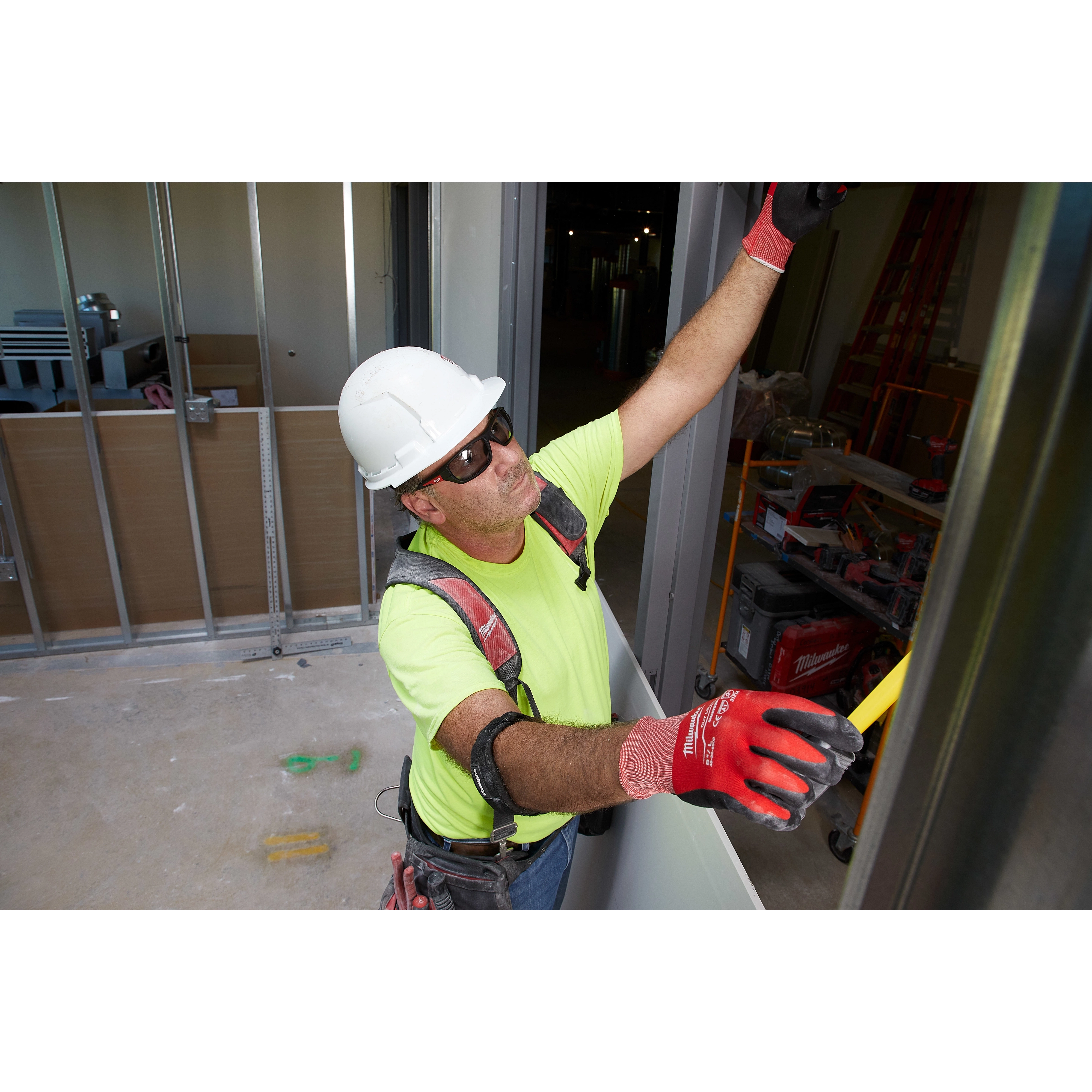 Construction worker wearing Safety Glasses - Gray Fog-Free Lenses, a hard hat, and safety harness installing drywall indoors.