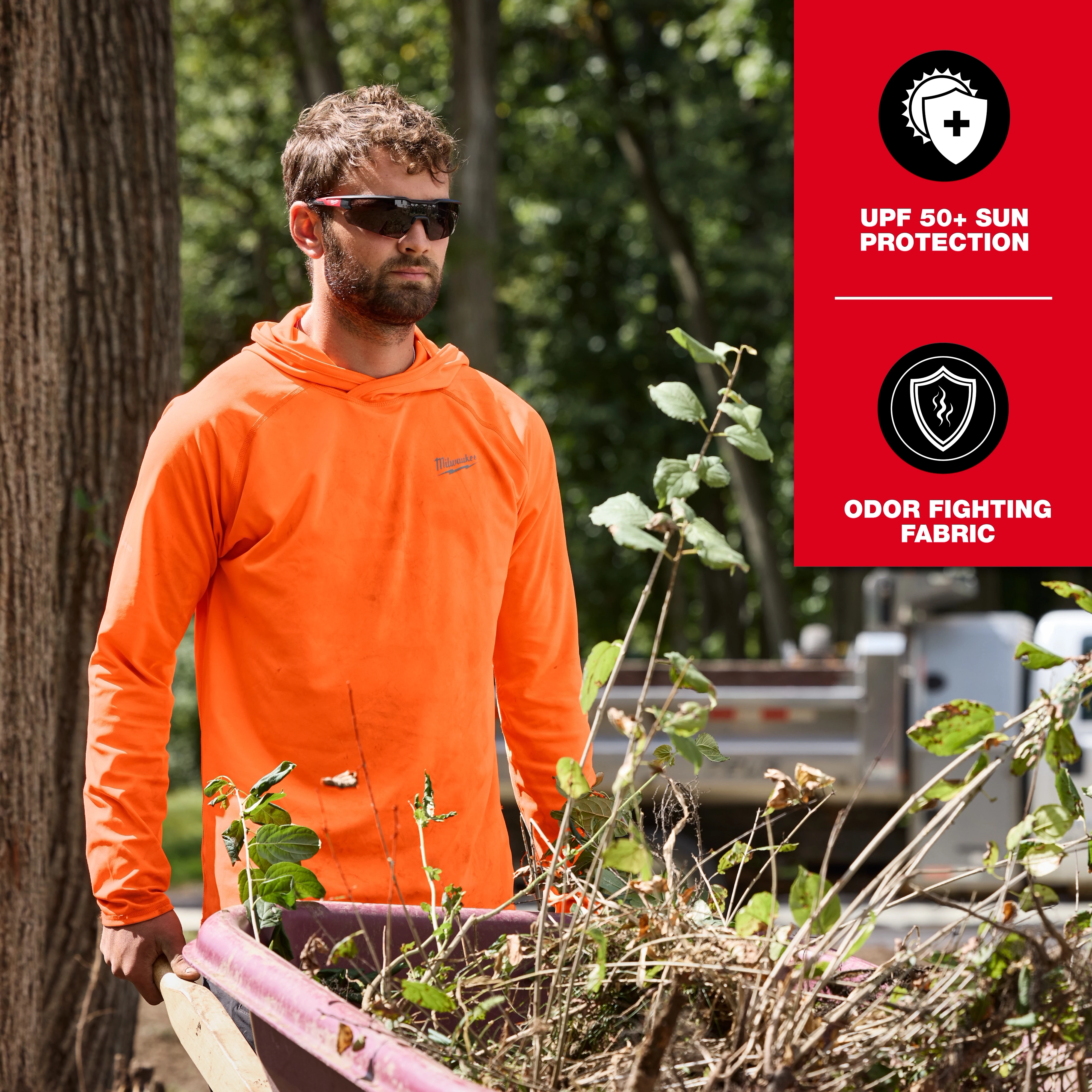 A person wearing an orange WORKSKIN Hooded Sun Shirt stands outdoors next to a wheelbarrow filled with branches. The shirt offers UPF 50+ sun protection and features odor-fighting fabric.