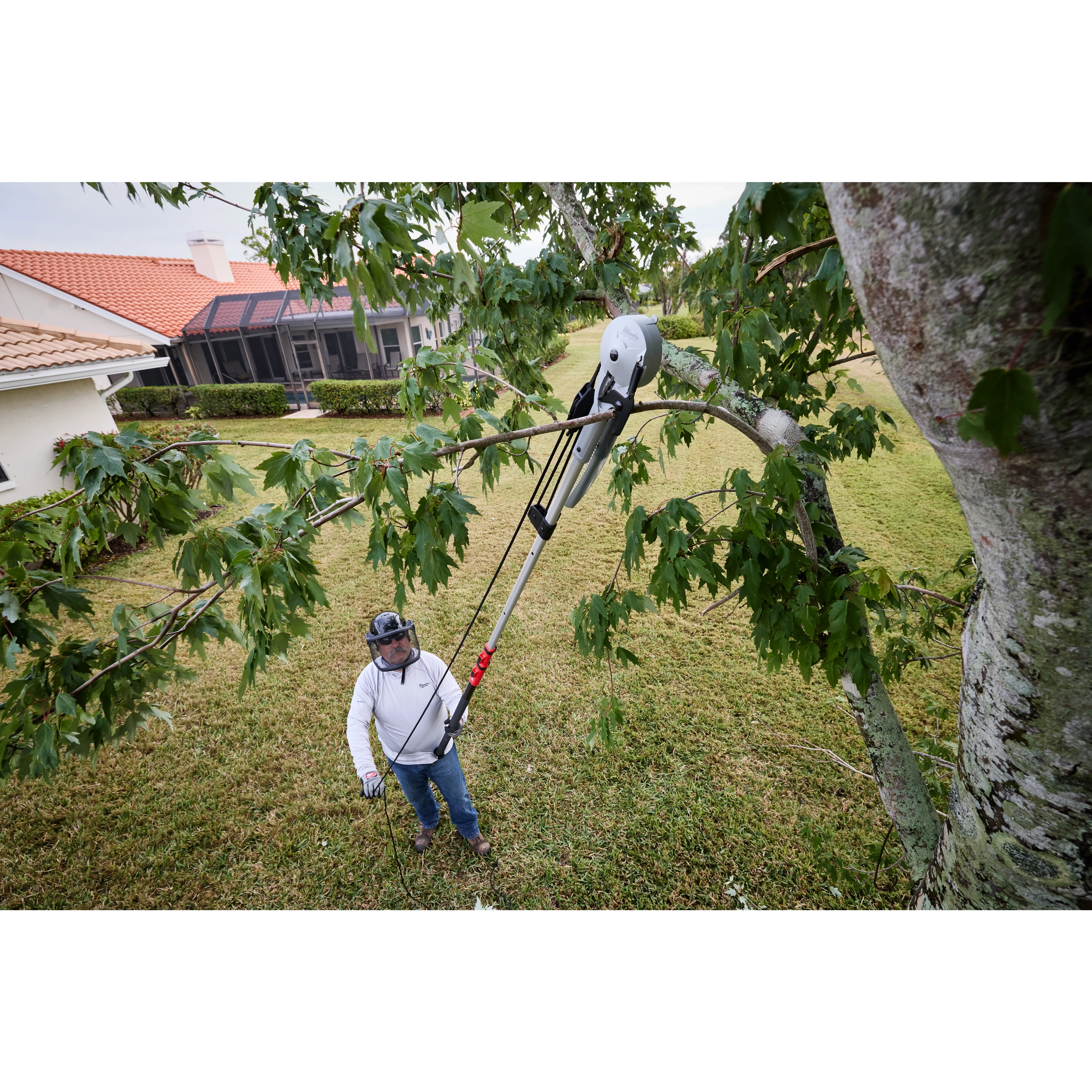 A person uses a 16' Pole Saw & Pruner to trim a tree branch in a yard. The tool is extended upward, cutting through a thick limb while the person stands on the ground. A house and lawn are visible in the background.