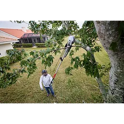 A person uses a 16' Pole Saw & Pruner to trim a tree branch in a yard. The tool is extended upward, cutting through a thick limb while the person stands on the ground. A house and lawn are visible in the background.