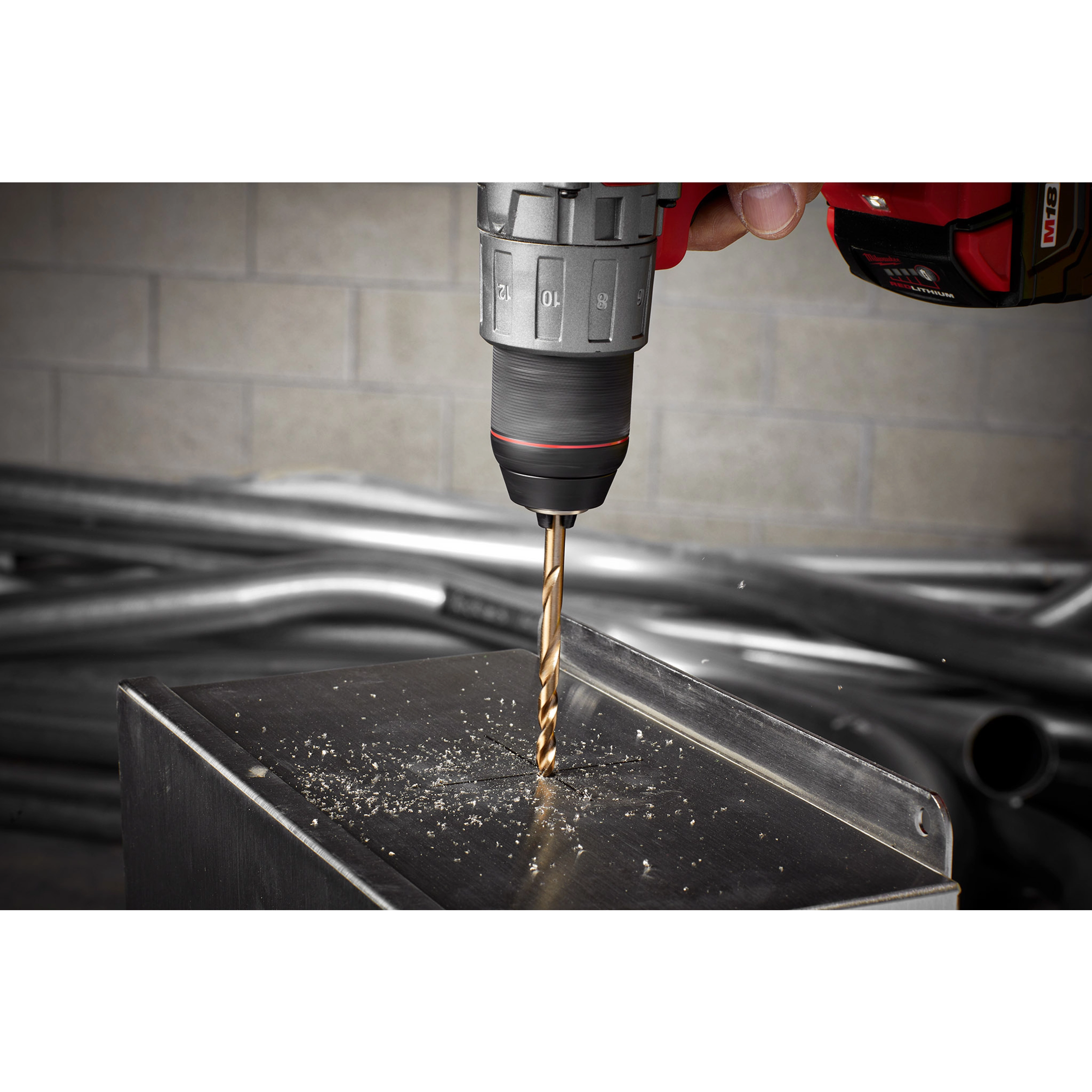 The RED HELIX™ Cobalt 3/8" Drill Bit is attached to a drill and is actively drilling into a metal surface, creating fine metal shavings. The drill is held by a hand and positioned above a square metal object, with industrial pipes in the background.