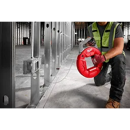 Person kneeling on a construction site holding a red 240' 1/8" Stainless Steel Fish Tape, threading wire through metal studs.
