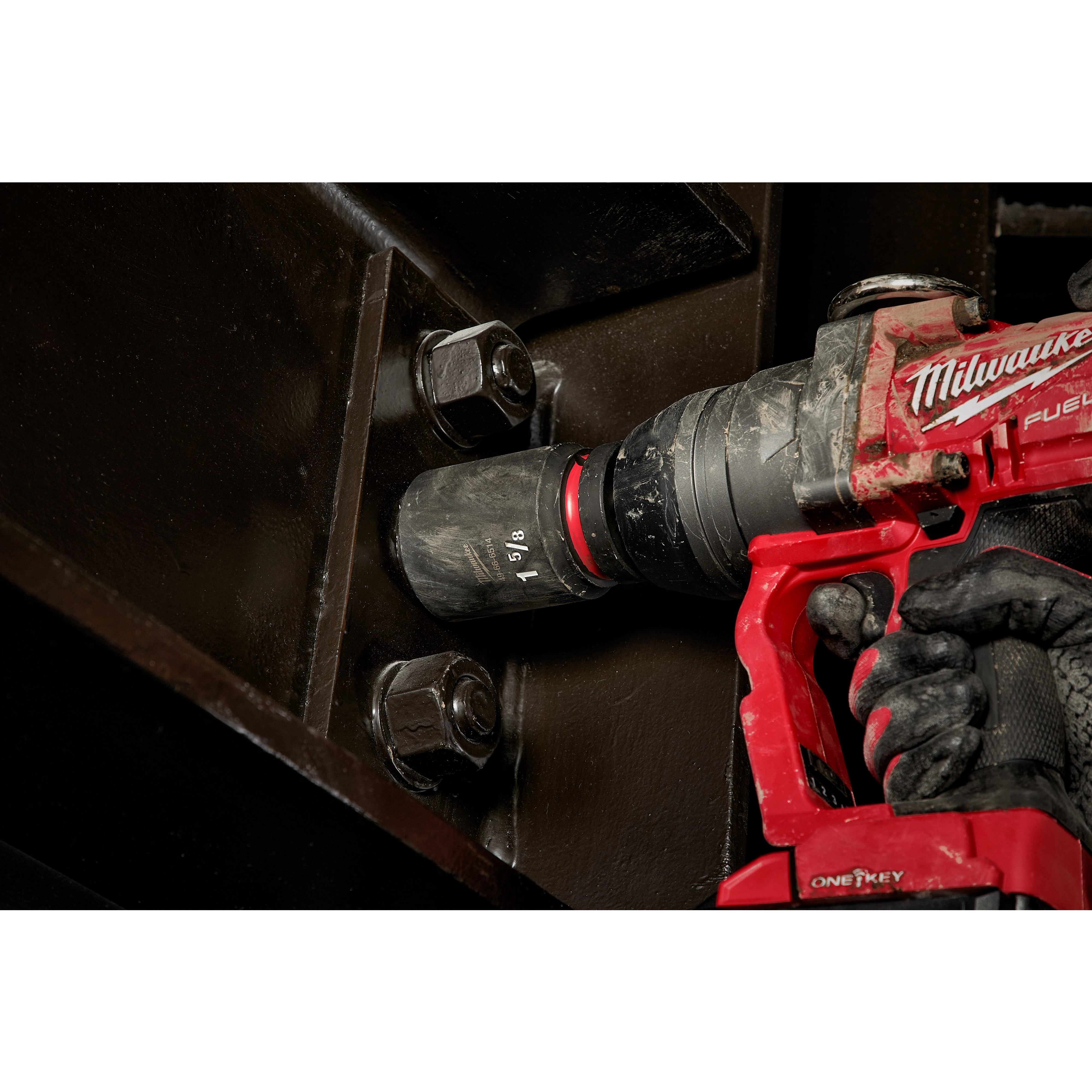 A red power tool is being used to tighten a large bolt with a SHOCKWAVE Impact Duty™ 1” Drive 1-5/8” Deep 6 Point Socket. The socket and tool are gripped by a gloved hand while engaging the bolt on a dark metal surface.