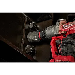 A red power tool is being used to tighten a large bolt with a SHOCKWAVE Impact Duty™ 1” Drive 1-5/8” Deep 6 Point Socket. The socket and tool are gripped by a gloved hand while engaging the bolt on a dark metal surface.