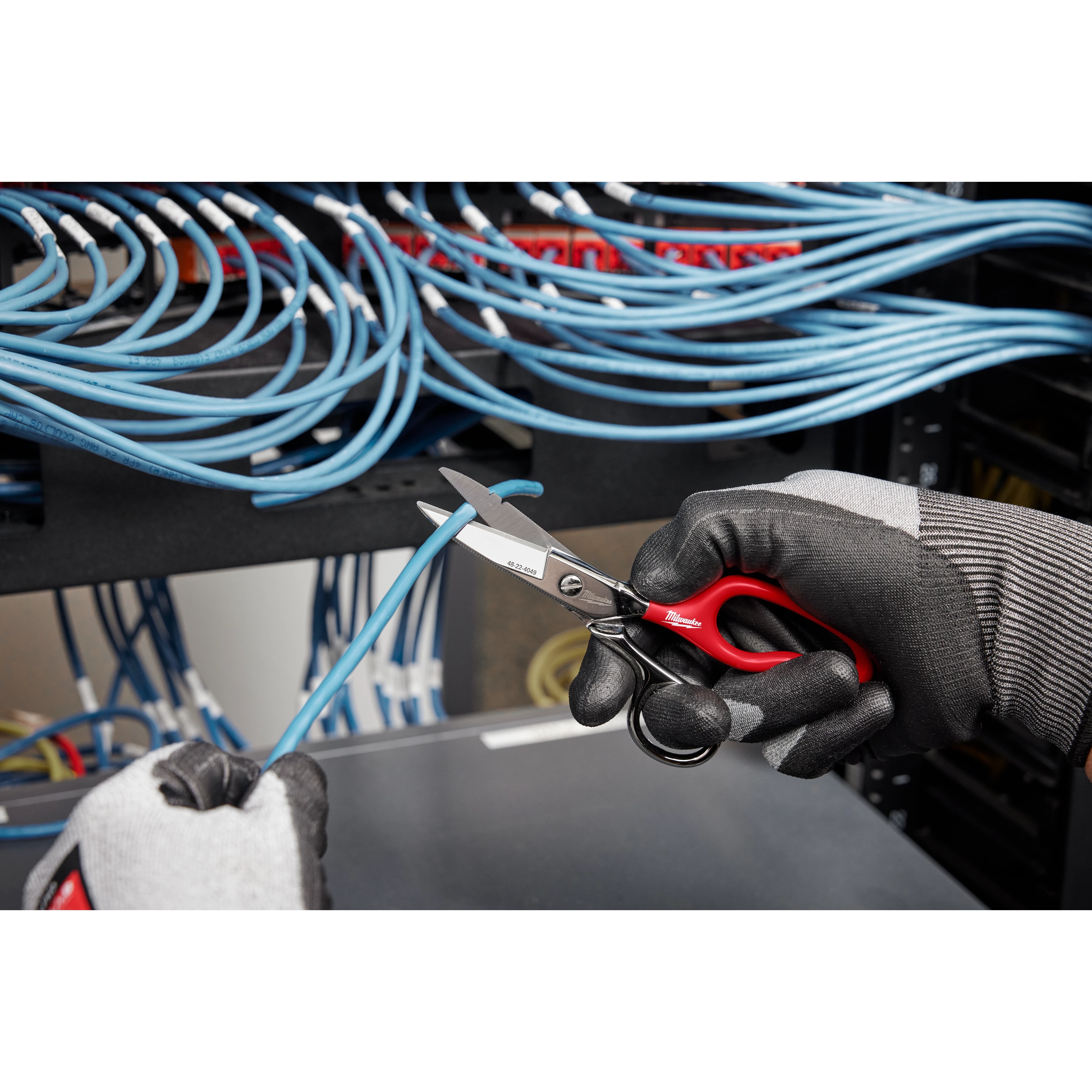 Electrician's gloved hands use Electrician Scissors with Extended Handle to cut a blue wire in a networking setup.