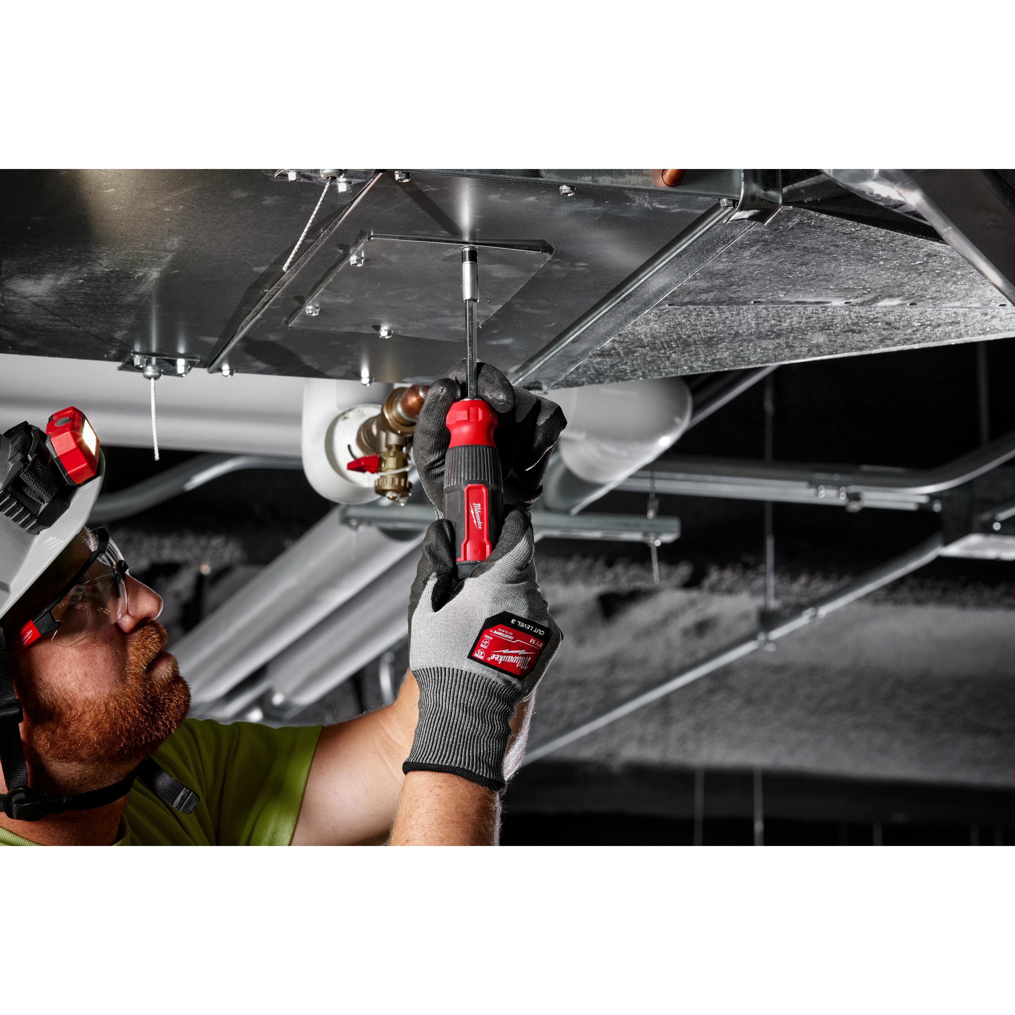 A person in a green shirt uses a 27-in-1 Multi-Bit Screwdriver to work on overhead metal ductwork in a building.