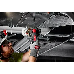 A person in a green shirt uses a 27-in-1 Multi-Bit Screwdriver to work on overhead metal ductwork in a building.