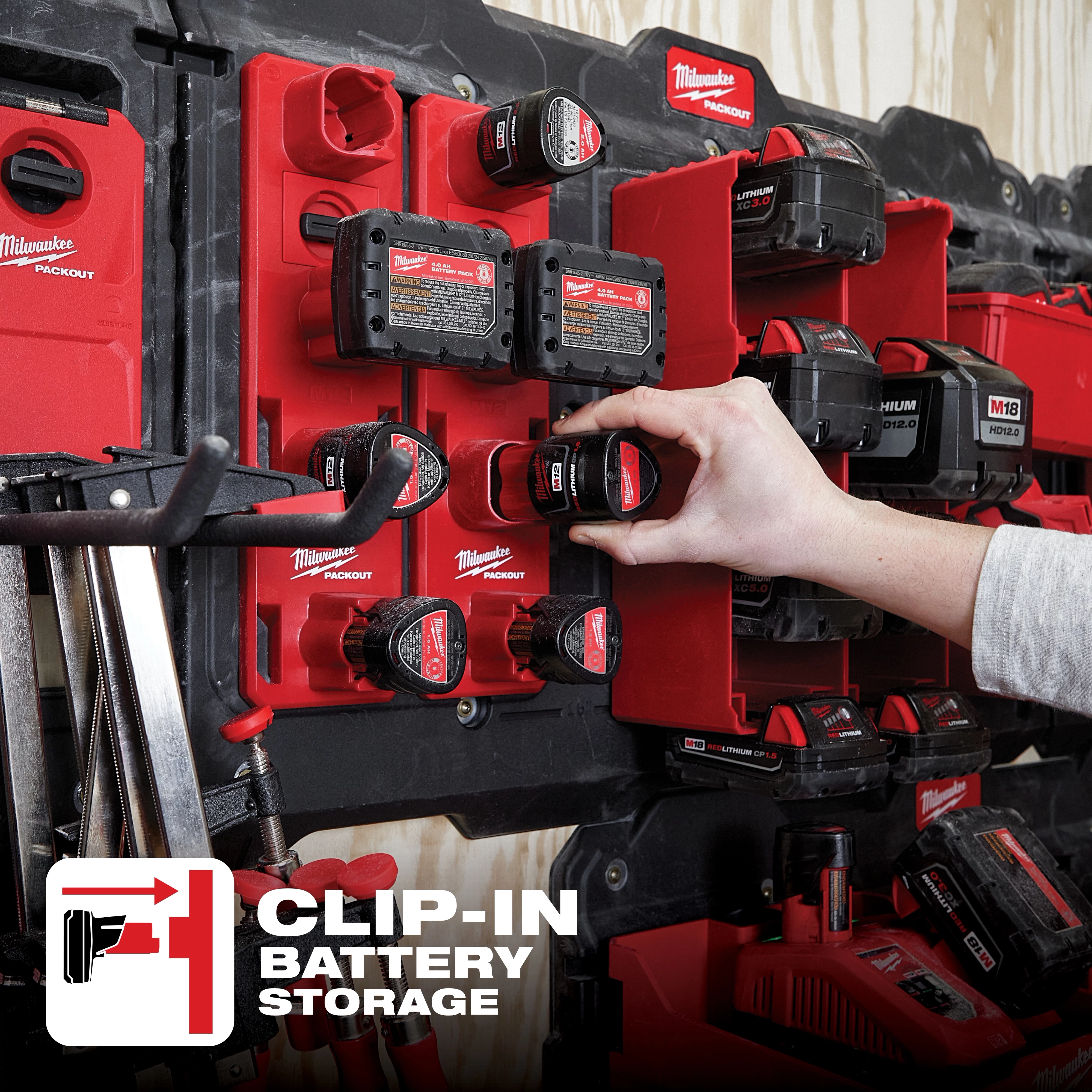 Hand places a battery in a PACKOUT M12 Battery Rack on a wall displaying multiple red battery packs and battery storage units.