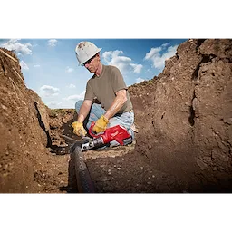 A worker uses the FORCELOGIC™ M18™ 3" Underground Cable Cutter Kit to cut a large underground cable. The tool is red and designed for heavy-duty industrial use. The worker is wearing yellow gloves, a white hard hat, and sits in a trench on a sunny day.