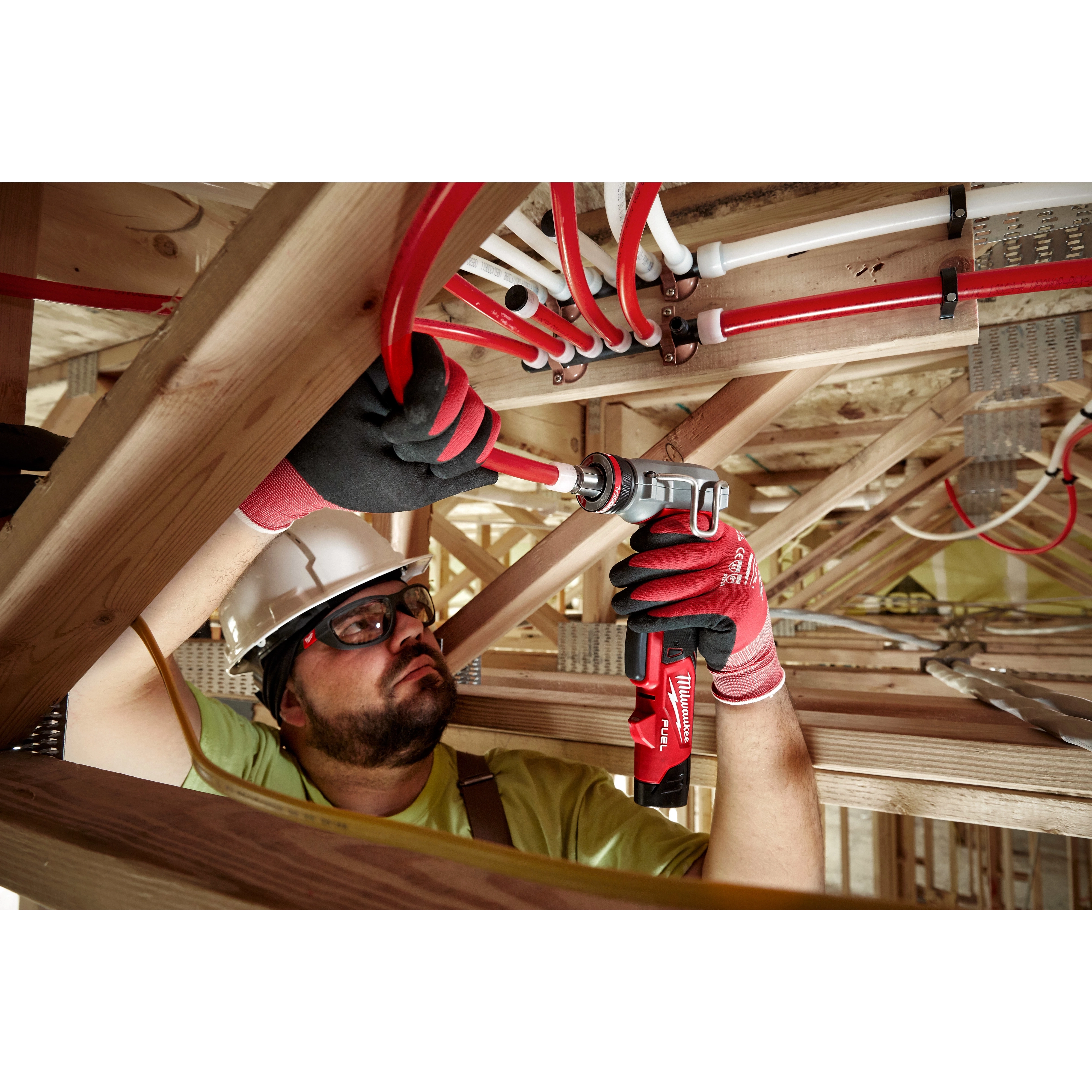 Man wearing gloves using M12 FUEL ProPEX Expander on red PEX pipes in wooden scaffolding.