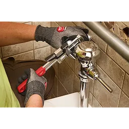 Gloved hands using a 12” Quick Adjust Smooth Jaw Wrench to tighten a metal flush valve in a tiled restroom.