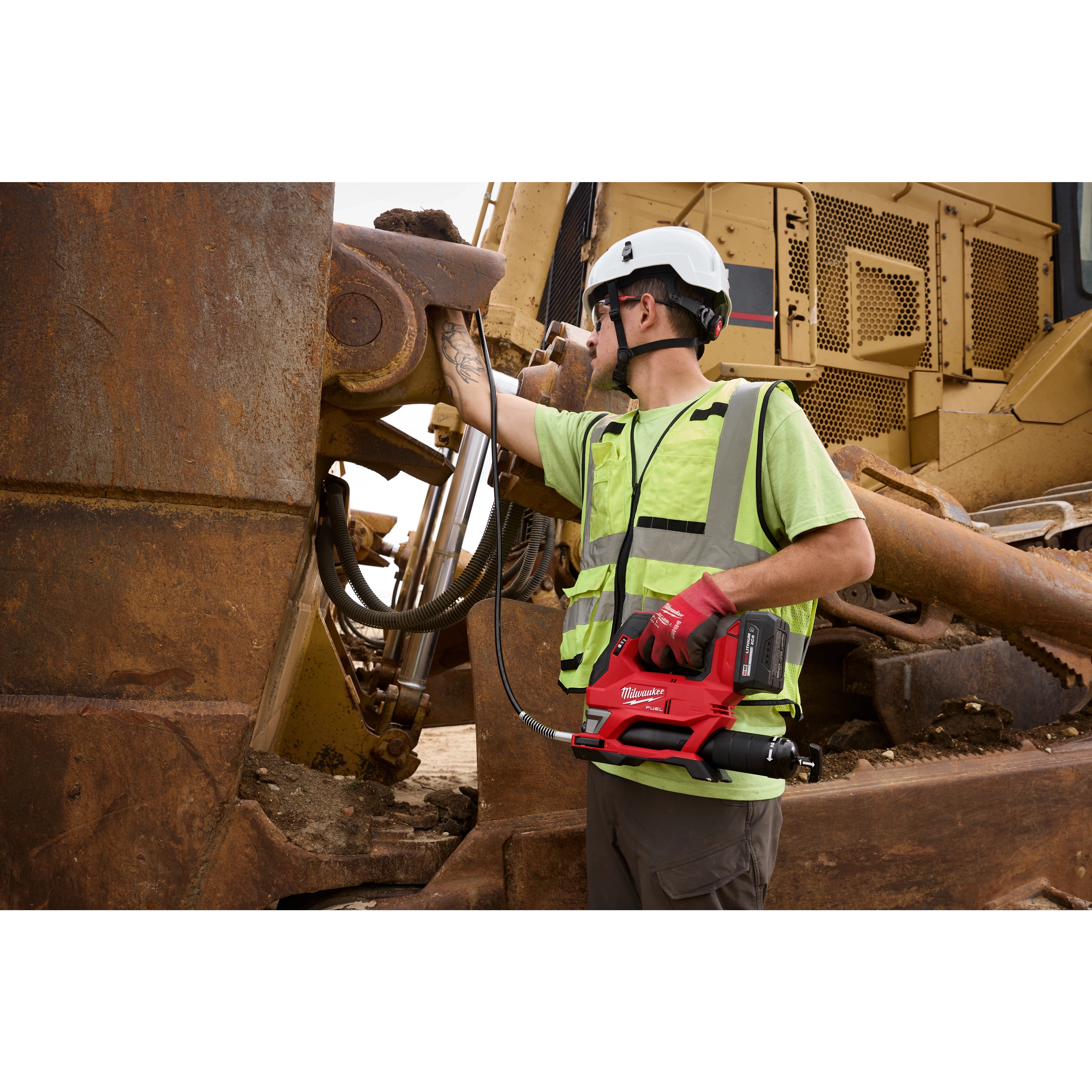 Worker in safety gear using an M18 FUEL Grease Gun on large rusted construction machinery.