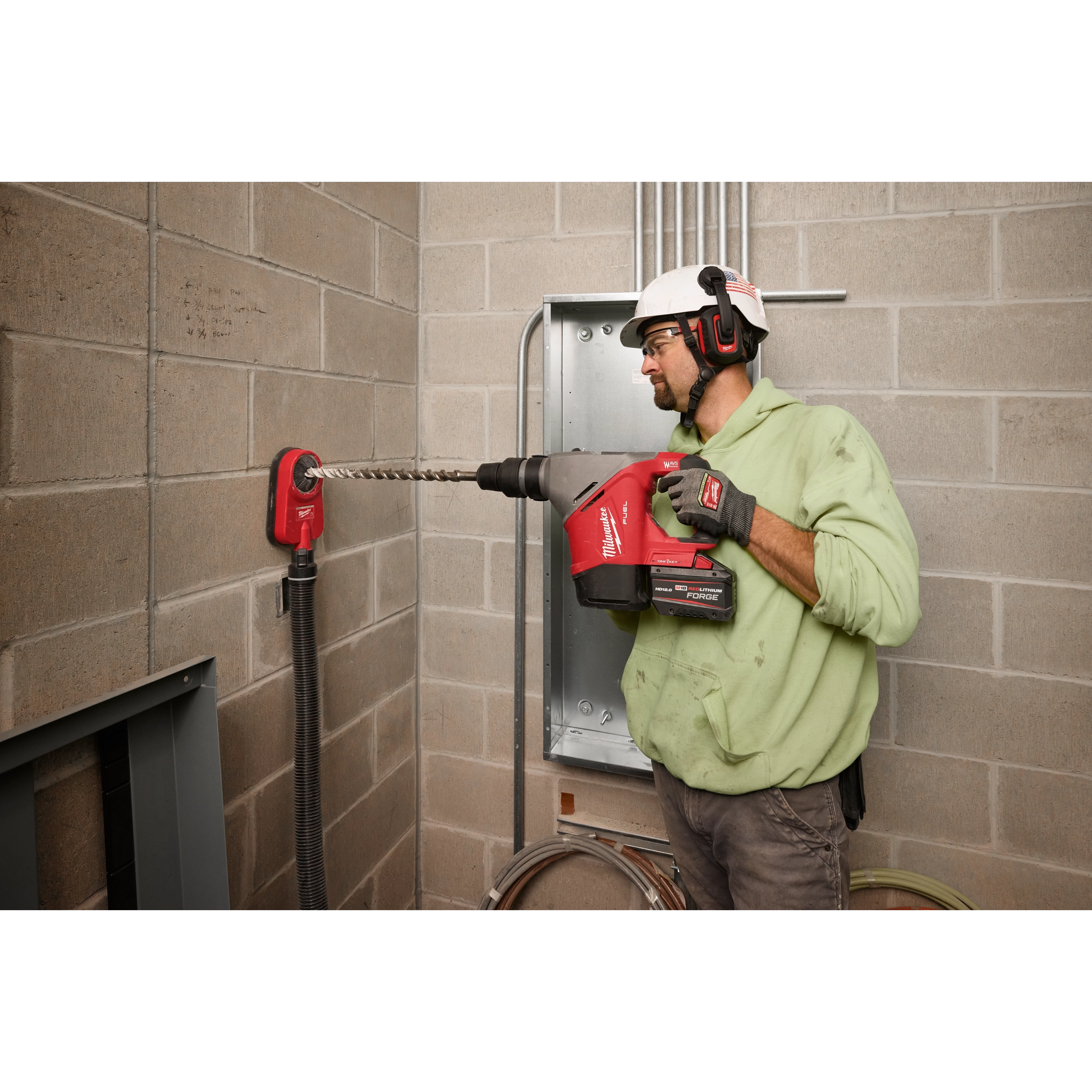 A person using the M18™ FUEL™ 1-9/16" SDS Max Rotary Hammer with ONE-KEY™ to drill into a concrete wall, wearing a hard hat and ear protection. The tool is red and connected to a dust extraction system.