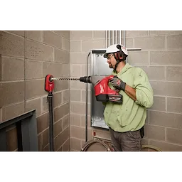 A person using the M18™ FUEL™ 1-9/16" SDS Max Rotary Hammer with ONE-KEY™ to drill into a concrete wall, wearing a hard hat and ear protection. The tool is red and connected to a dust extraction system.