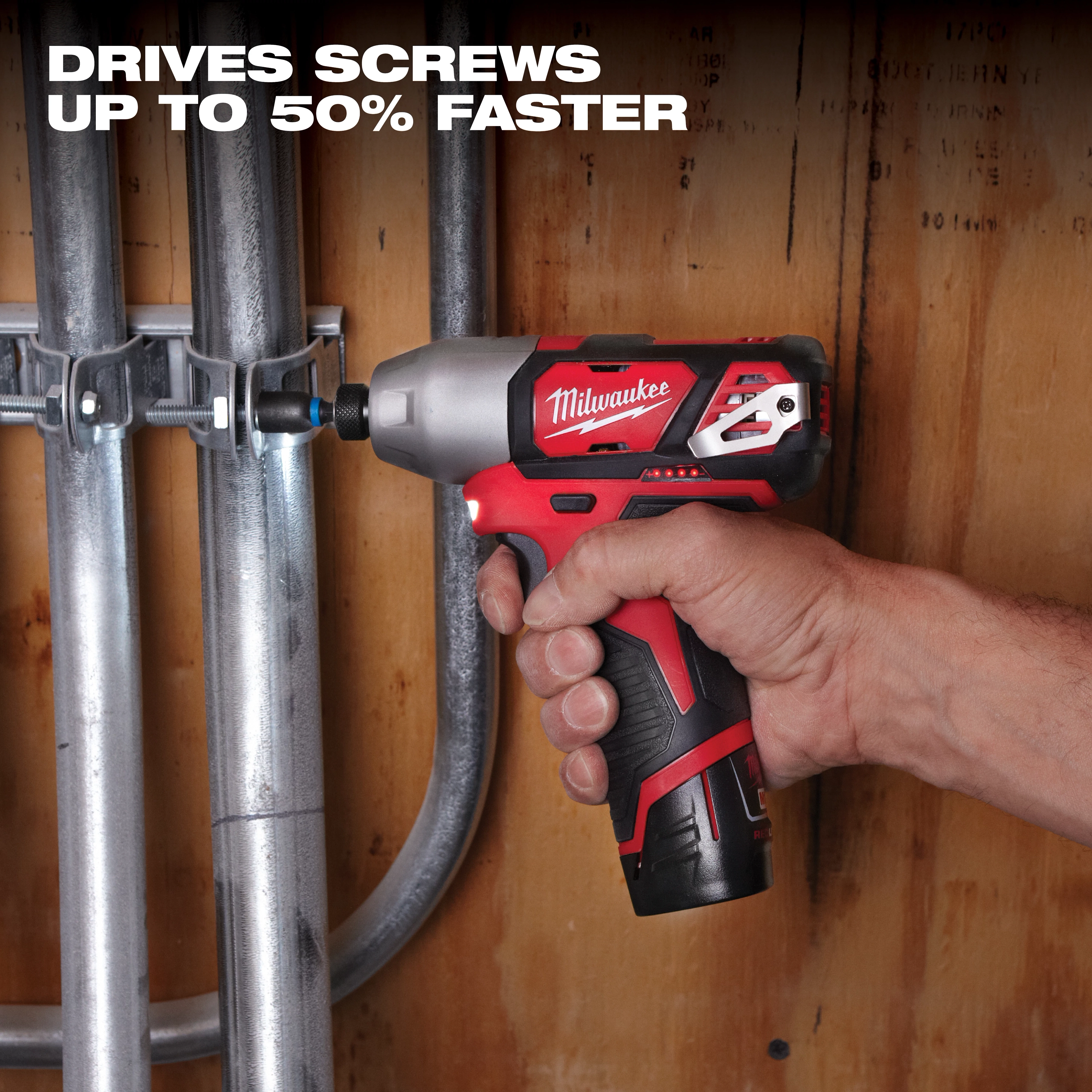 A person uses a red and black power driver tool from the M12 Cordless 4-Tool Combo Kit to fasten a screw to secure metal pipes against a wooden wall. Text above reads, "DRIVES SCREWS UP TO 50% FASTER."